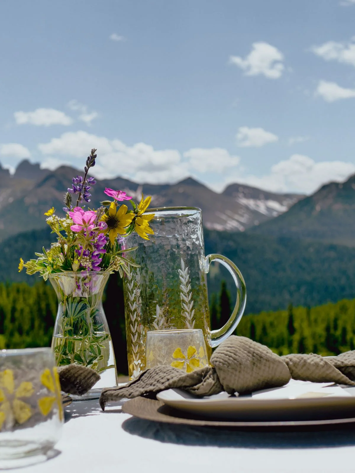 Table with a glass pitcher, small glass, and a vase with colorful wildflowers, set outdoors with a mountain landscape and blue sky in the background.