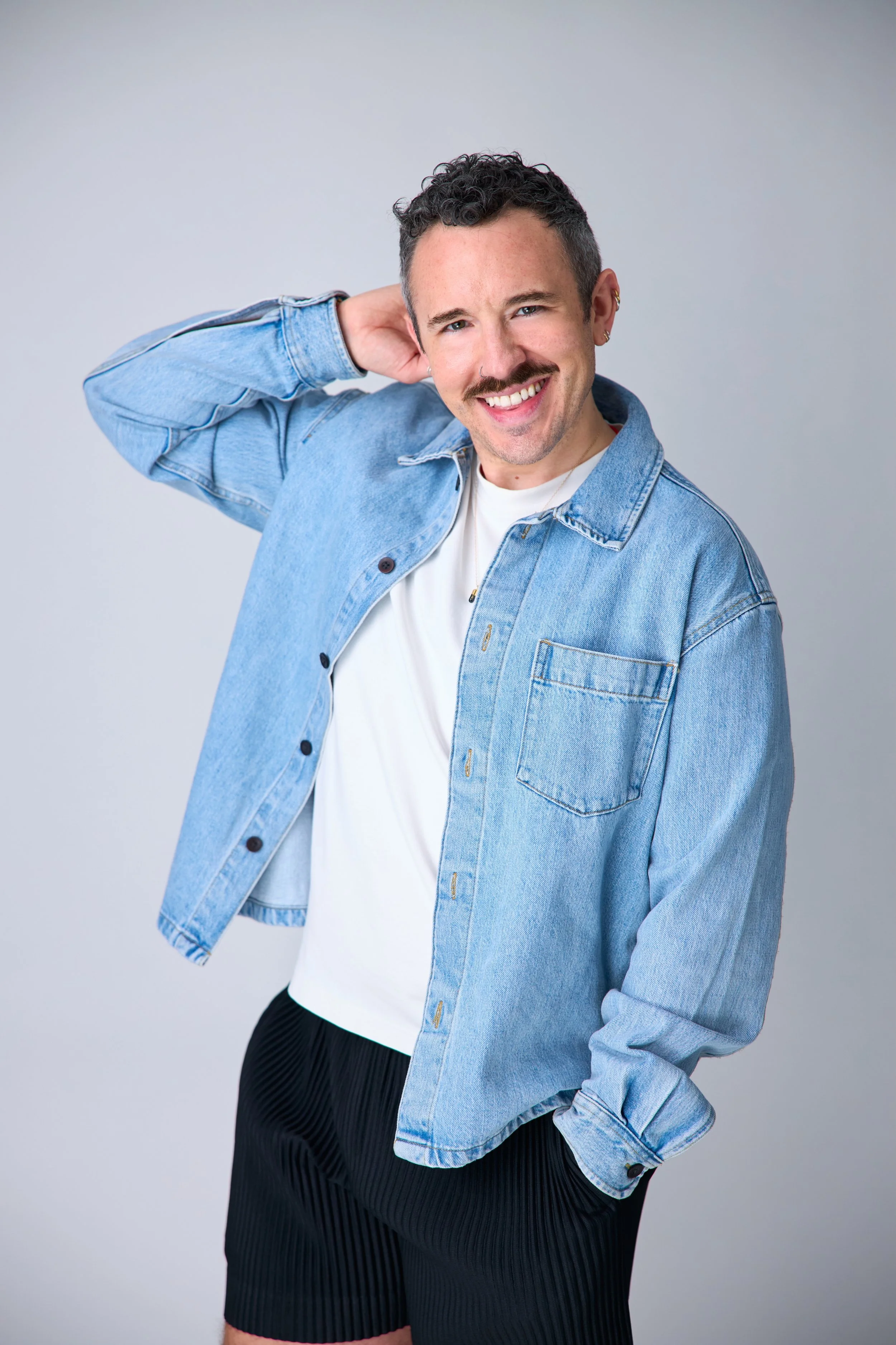 A young man with curly dark hair and a mustache smiling, wearing a light blue denim jacket over a white t-shirt, black shorts, and standing against a plain gray background.