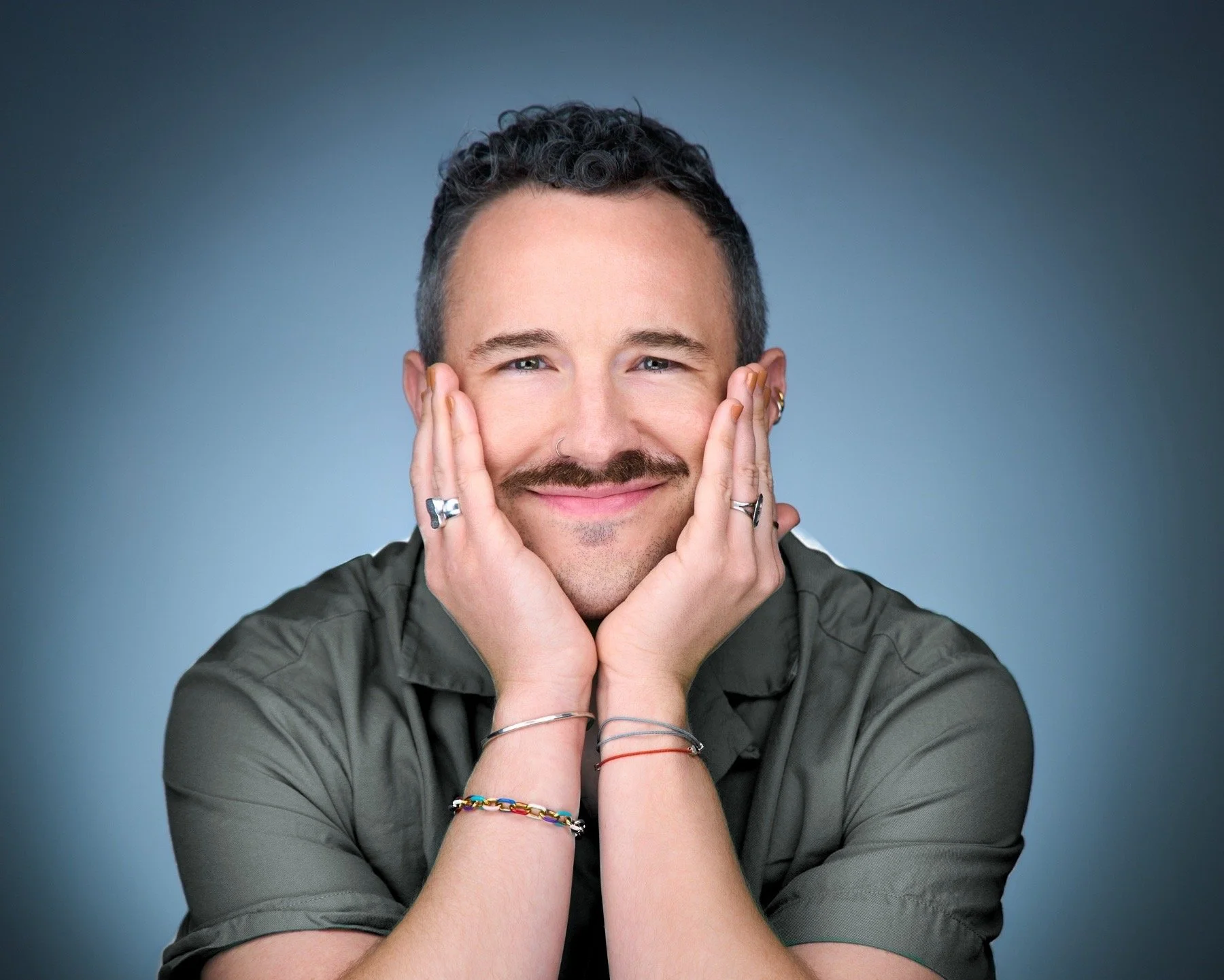 A smiling man with short dark curly hair, a mustache, and wearing multiple rings and bracelets, resting his face in his hands against a gradient blue background.