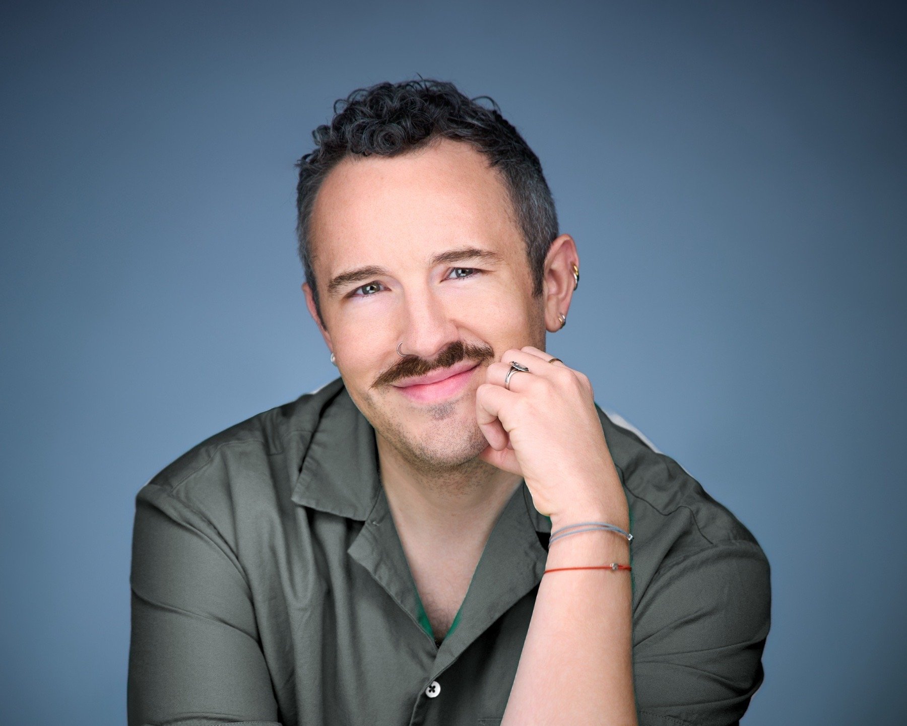 A man with curly dark hair, a mustache, and light skin, smiling at the camera, resting his chin on his hand, against a blue background.