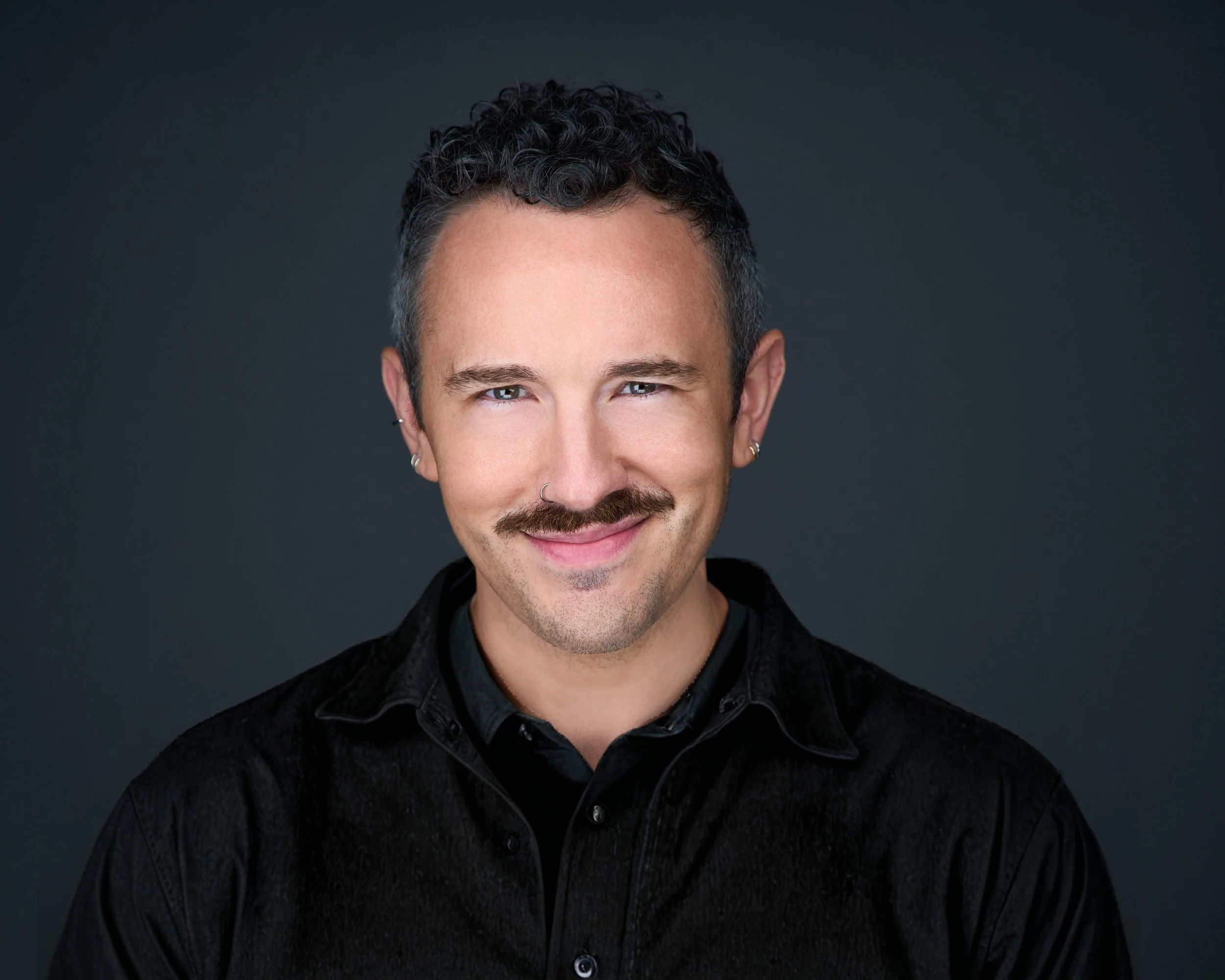 Portrait of a man with short curly dark hair, light skin, blue eyes, mustache, beard, and earrings, smiling against a dark background.
