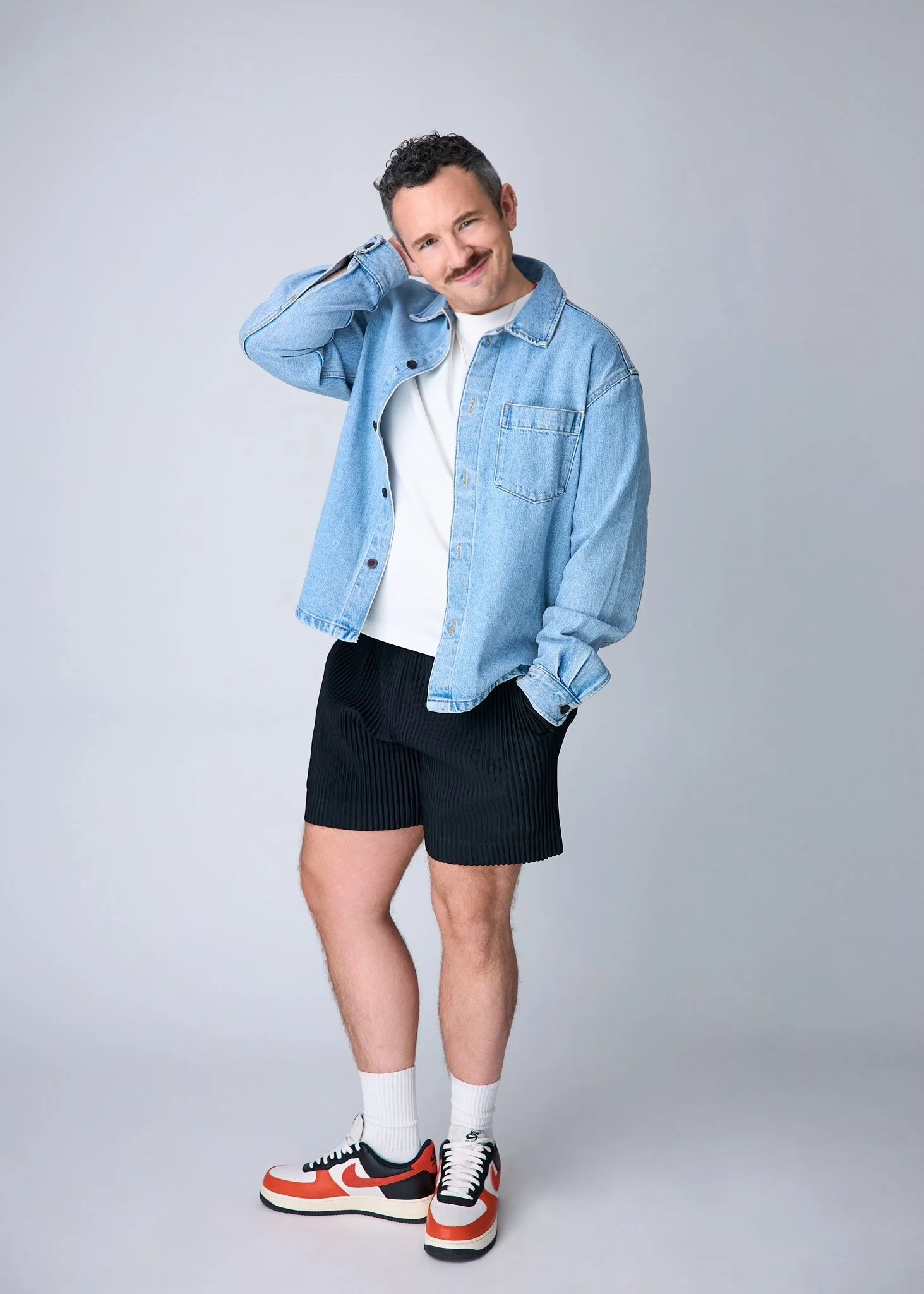 A man wearing a light blue denim jacket, white T-shirt, black pleated shorts, white socks, and Nike sneakers posing against a plain background.