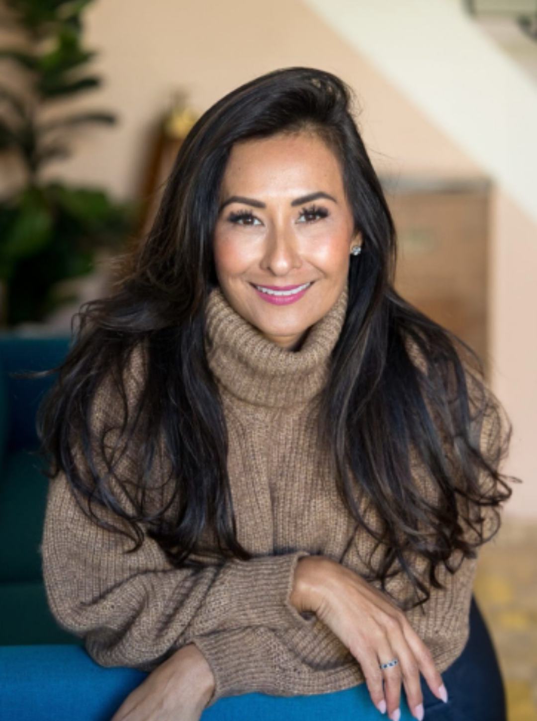 A woman with long black hair, wearing a tan turtleneck sweater, smiling while sitting indoors.