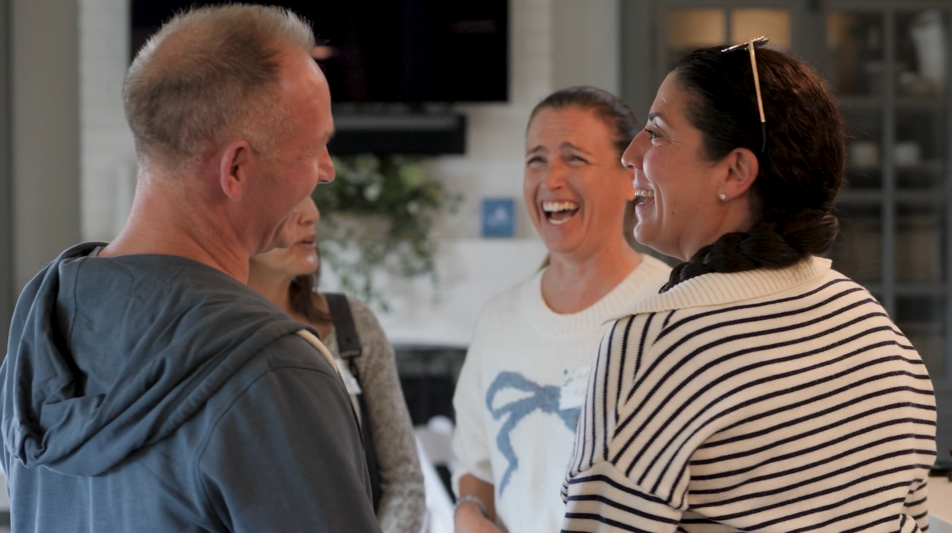 Four people laughing and talking indoors, three women, one man, casual clothing.