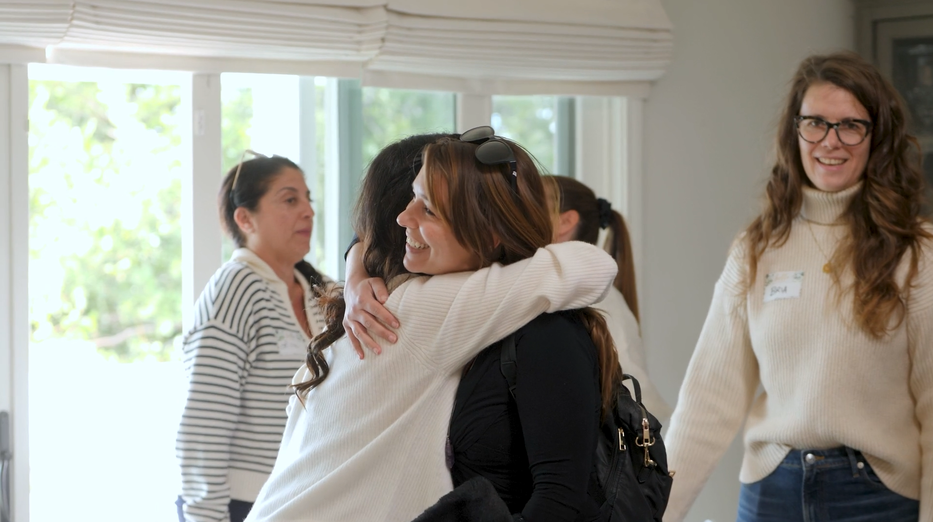 Two women hugging and smiling at a social gathering with others in the background.