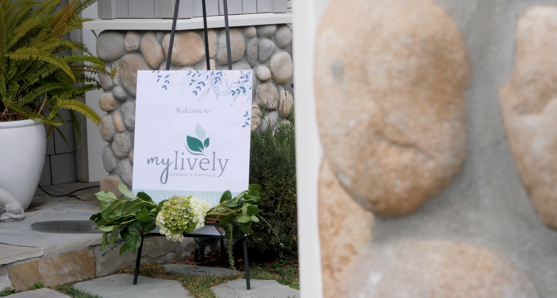 A welcome sign for 'mylively' with a leafy logo, placed outdoors near a stone wall and plants, with a floral arrangement in front.