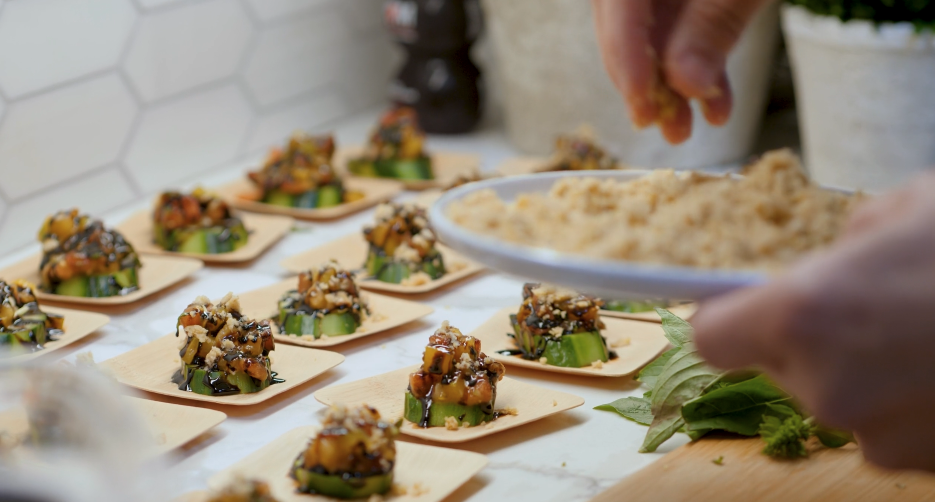 Person sprinkling topping onto small, sliced cucumber appetizers arranged on wooden trays.