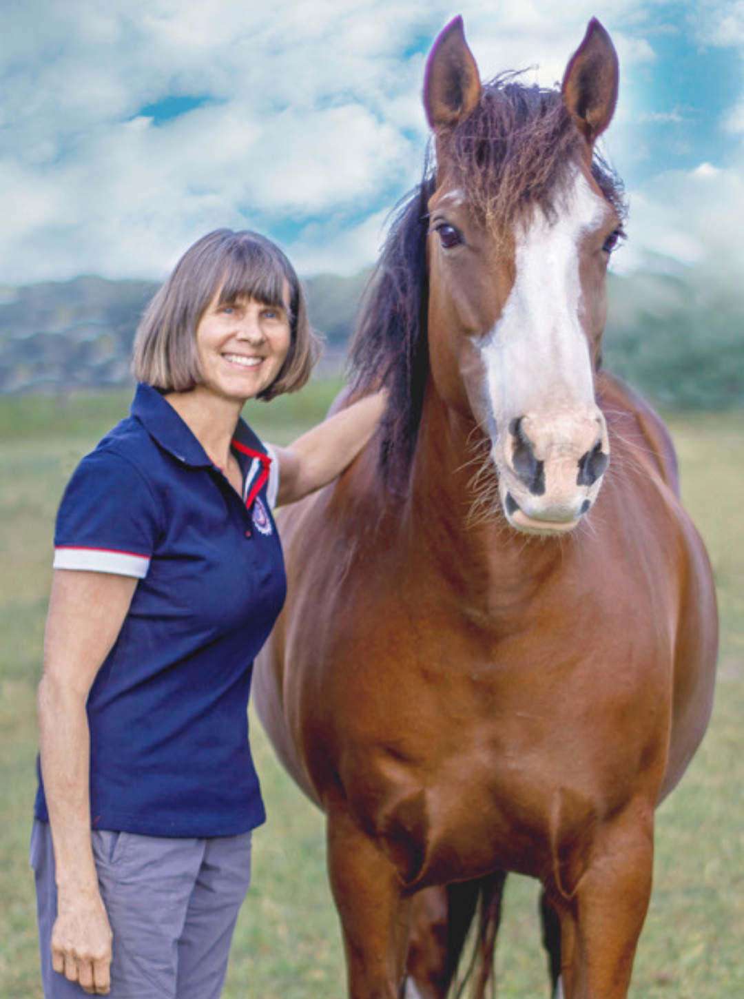 A woman with short brown hair smiling and wearing a navy polo shirt standing next to a large brown horse on a grassy field with a cloudy sky in the background.