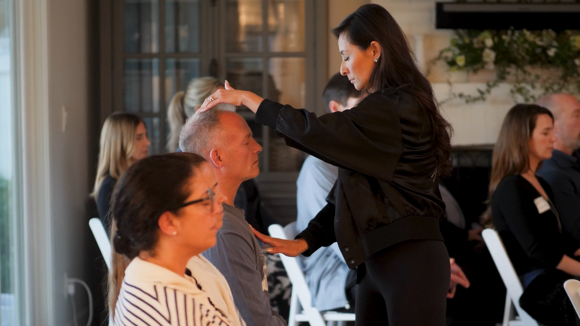 A woman with long dark hair, dressed in black, is praying over a seated man with short hair and a gray shirt. She has her hand on his head and is holding his shoulder. The room has several people sitting with their eyes closed, in a prayer or meditation setting.