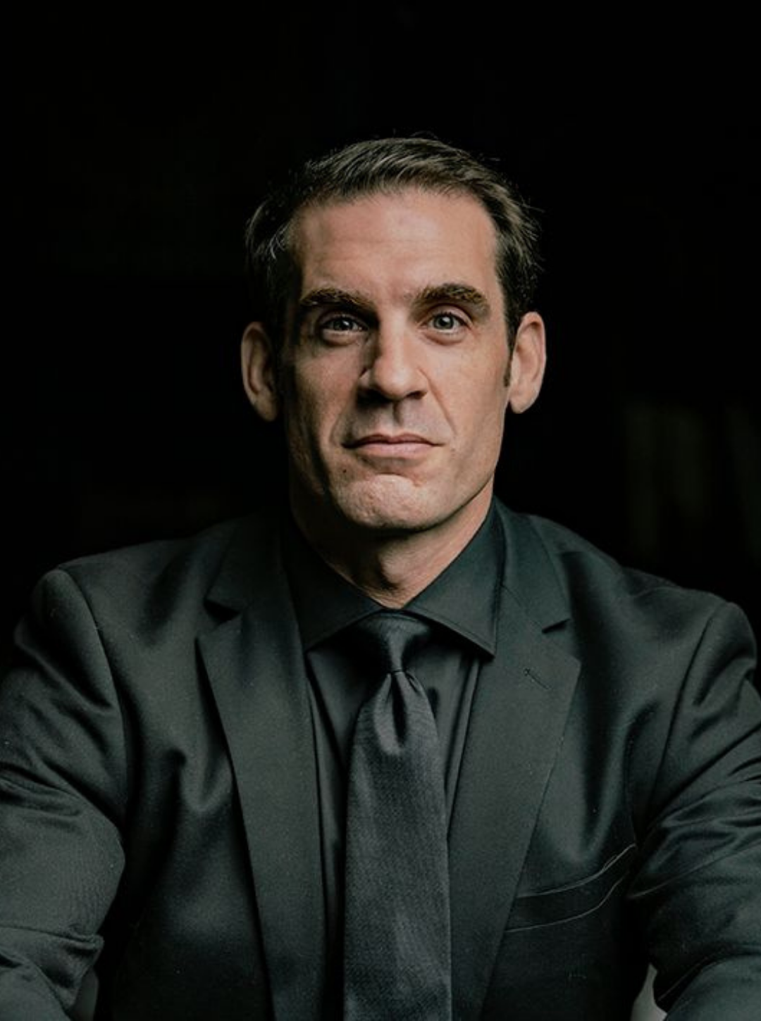 A man with dark hair in a black shirt and tie, sitting against a dark background.
