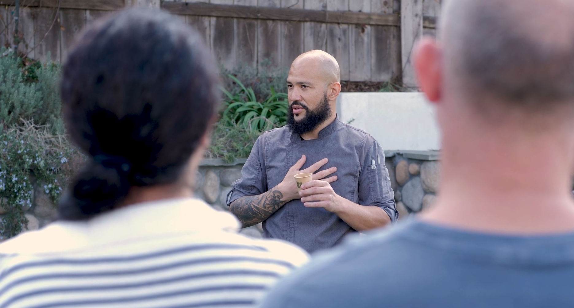 A man with a bald head and beard holding a small paper cup, speaking to a group outdoors with a wooden fence and plants in the background.