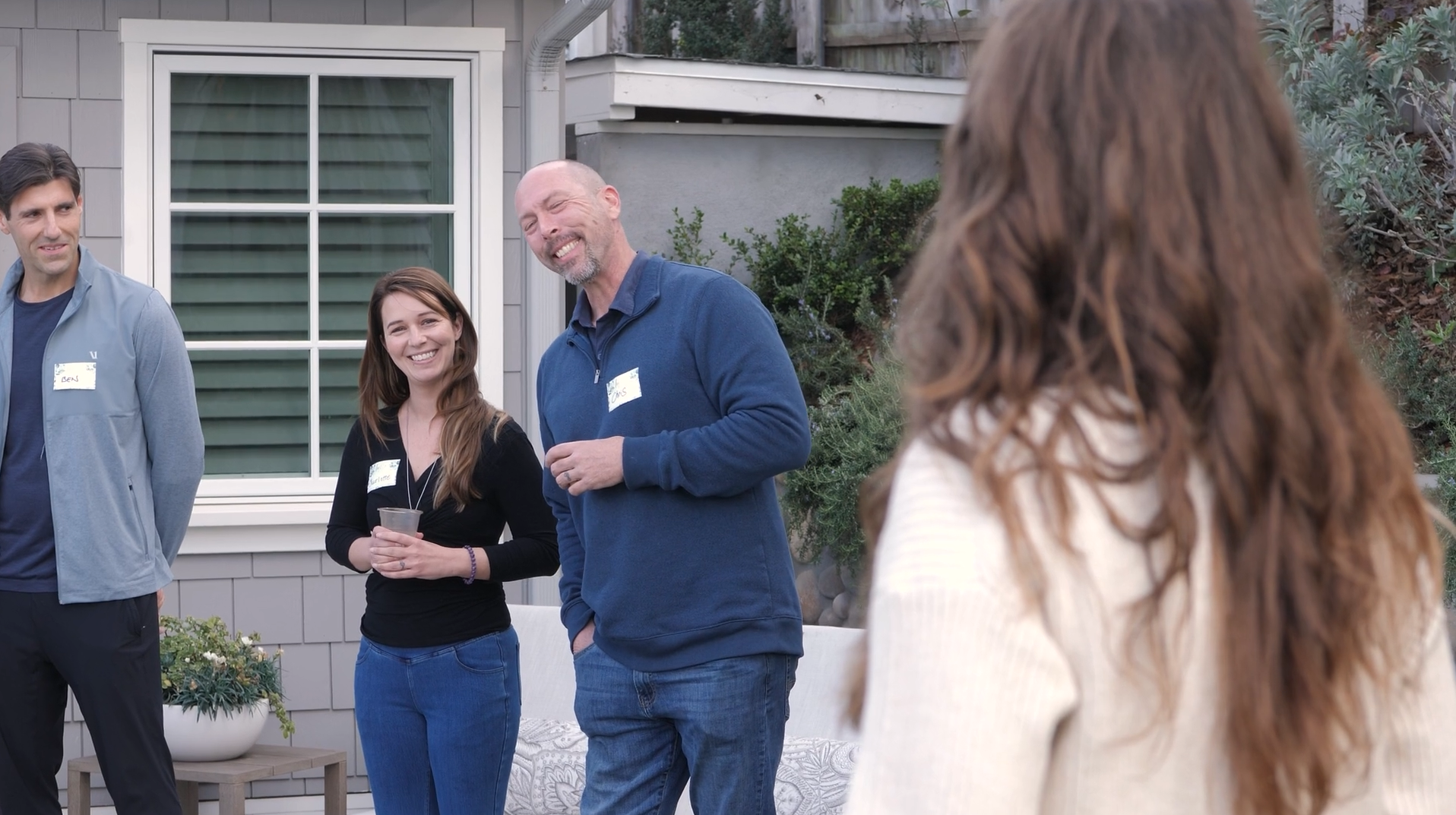 Four people at an outdoor gathering, three men and one woman, smiling and talking. The woman is holding a clear cup. They are standing on a porch or backyard area with plants and a house in the background.