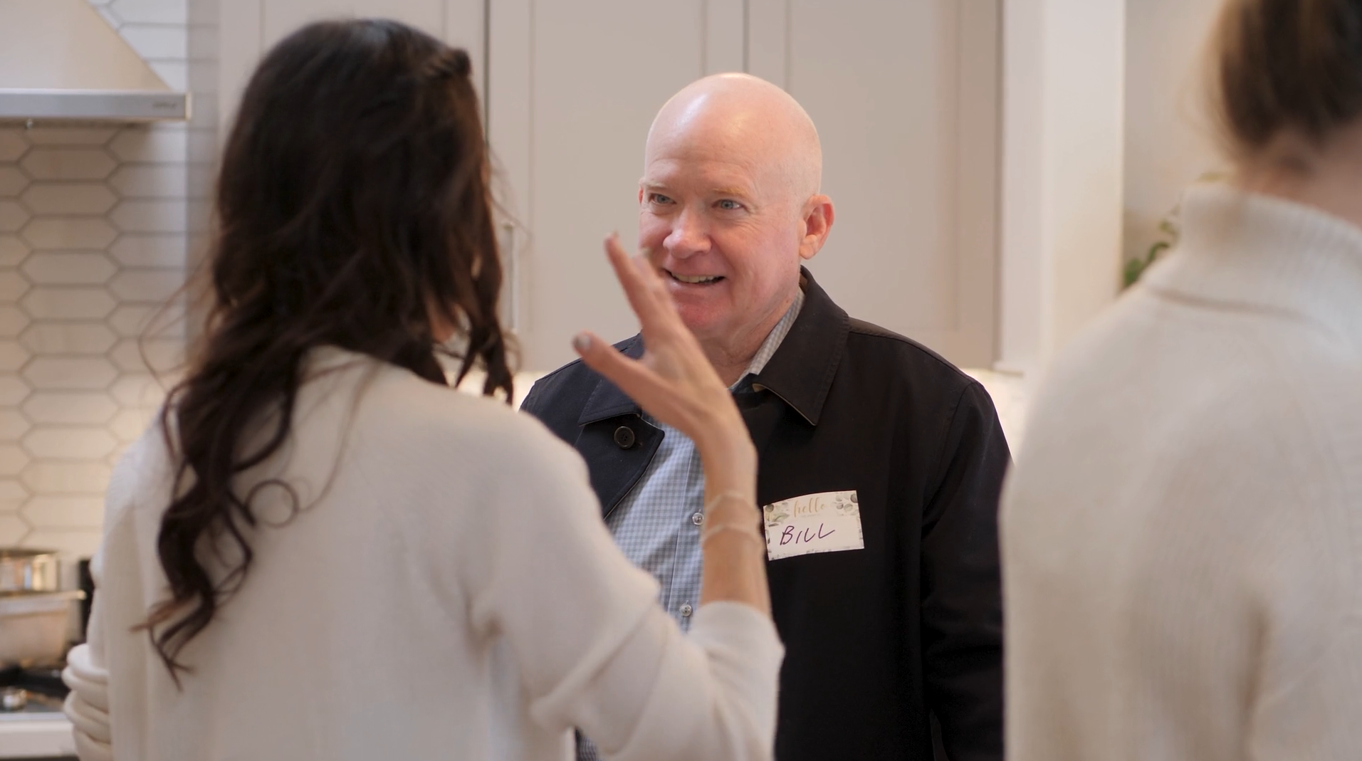 A man with a bald head and wearing a name tag that says 'Bill' is smiling and talking to two women in a kitchen.