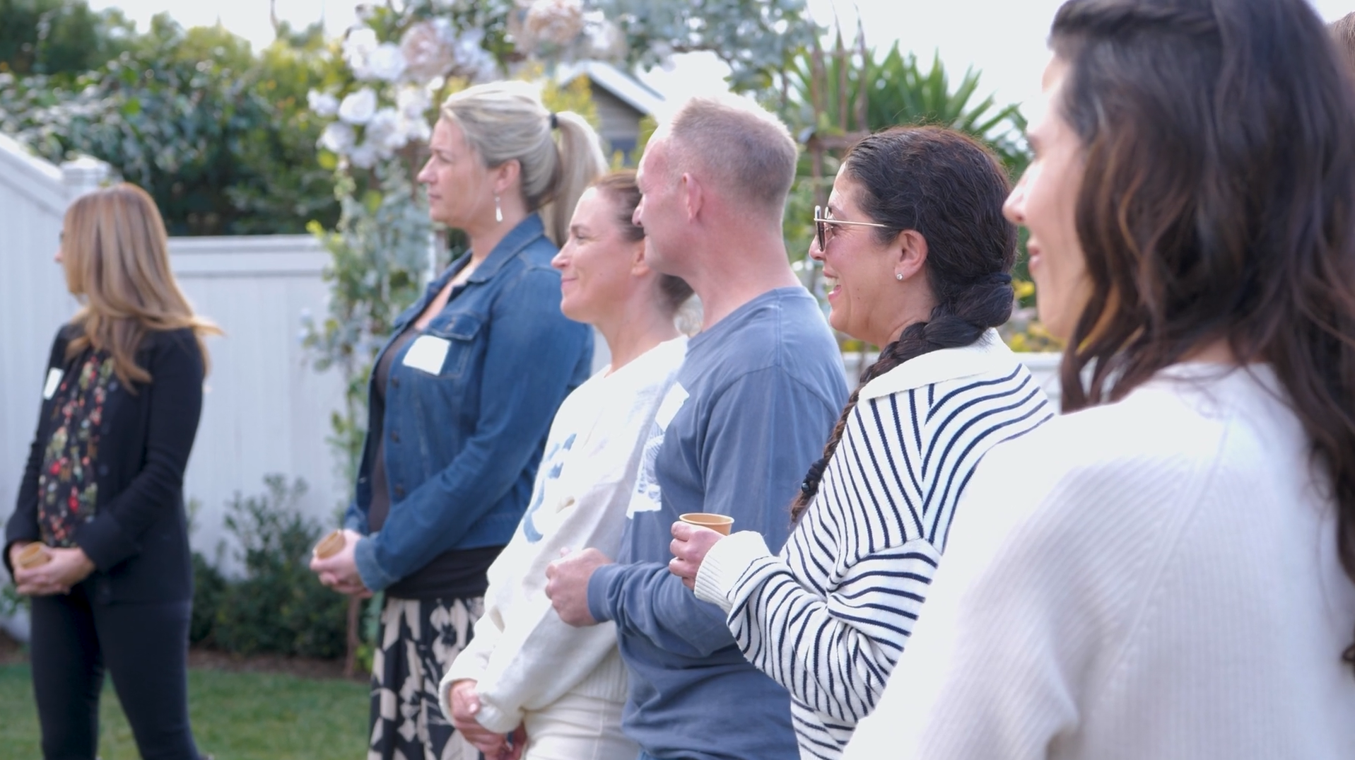 A group of people standing outdoors in a line, attending a social event or gathering, with some holding cups and smiling.