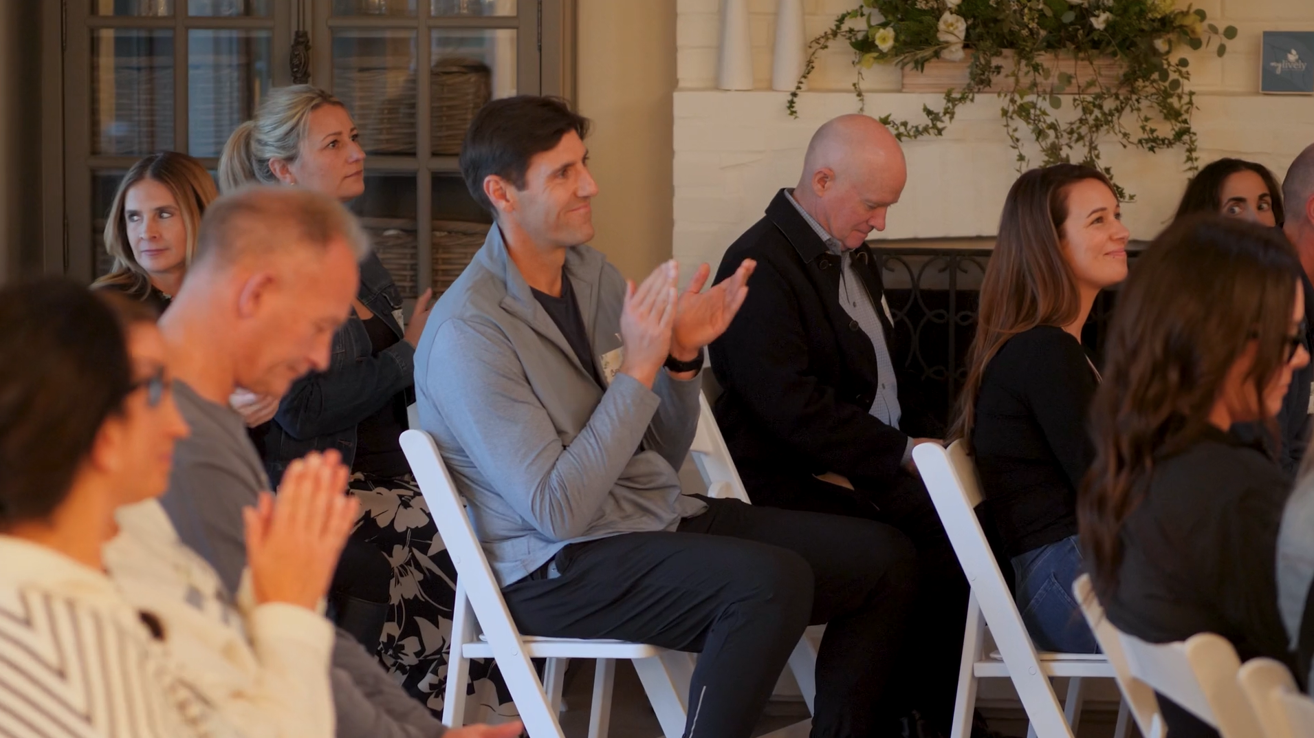People attending an indoor event, sitting on white chairs, some clapping, others with hands folded, in a warm, decorated space with a fireplace and plants.