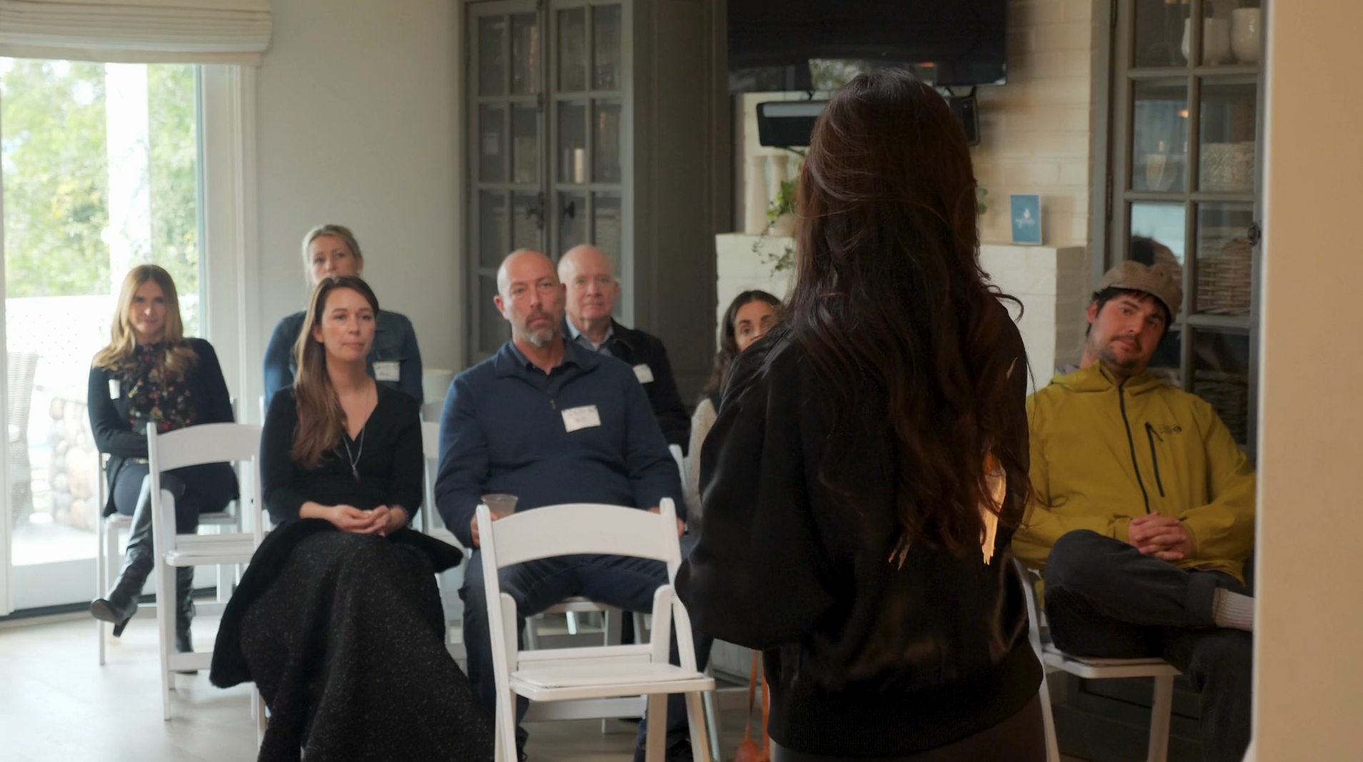 A woman with long dark hair speaking in front of an audience in a room with a large window and a flat-screen TV.