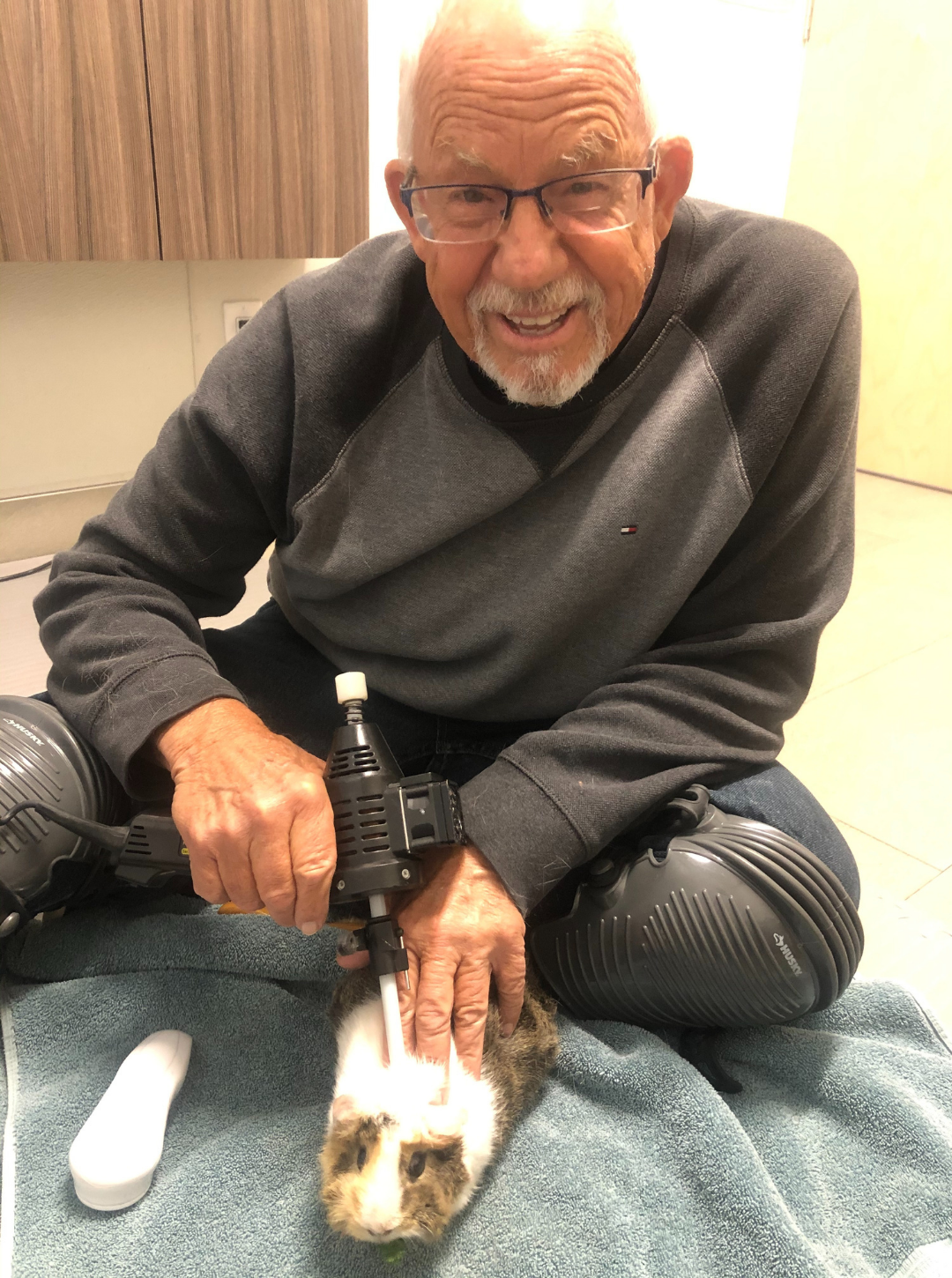 An elderly man with glasses smiling while holding a small guinea pig on a towel, with a handheld medical device nearby.