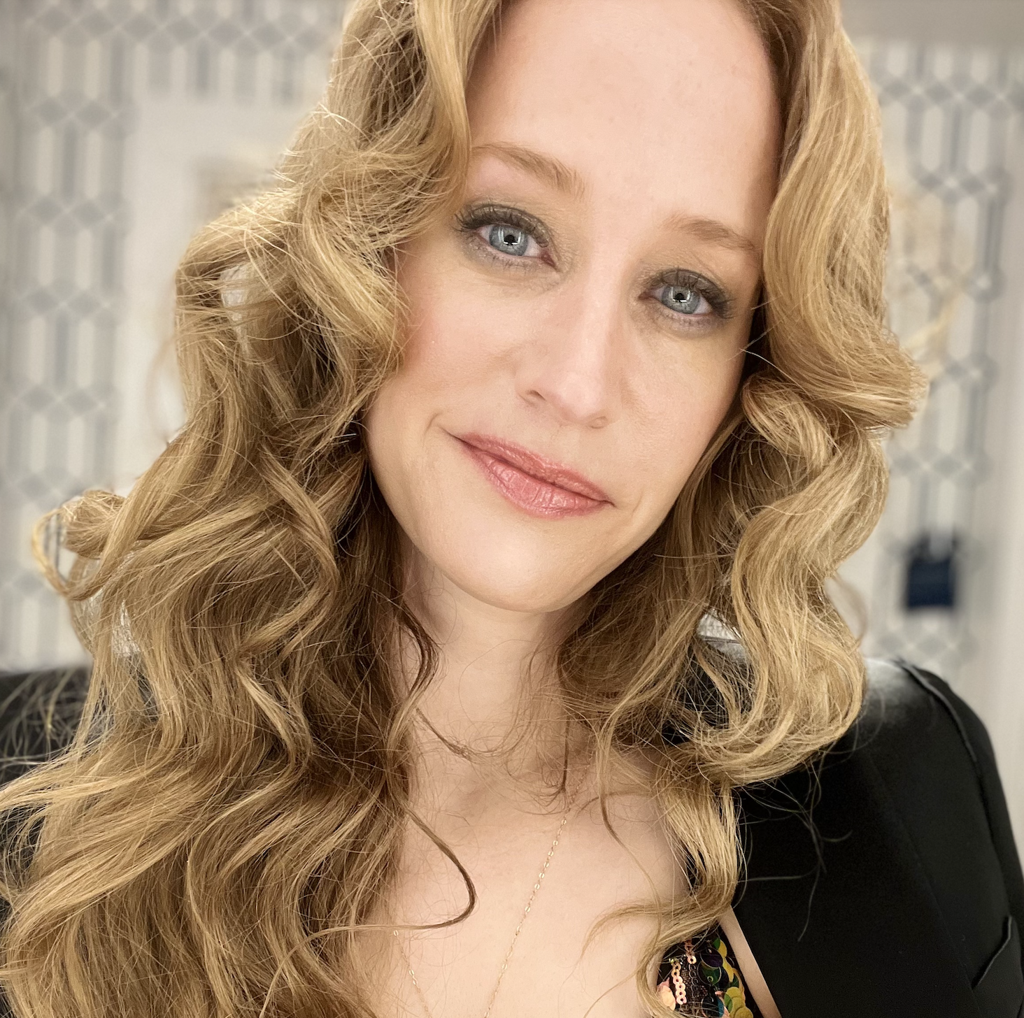 Close-up of a woman with long, wavy blonde hair, fair skin, and blue eyes, smiling softly, wearing a black top with a visible sequin detail, and a delicate gold necklace.