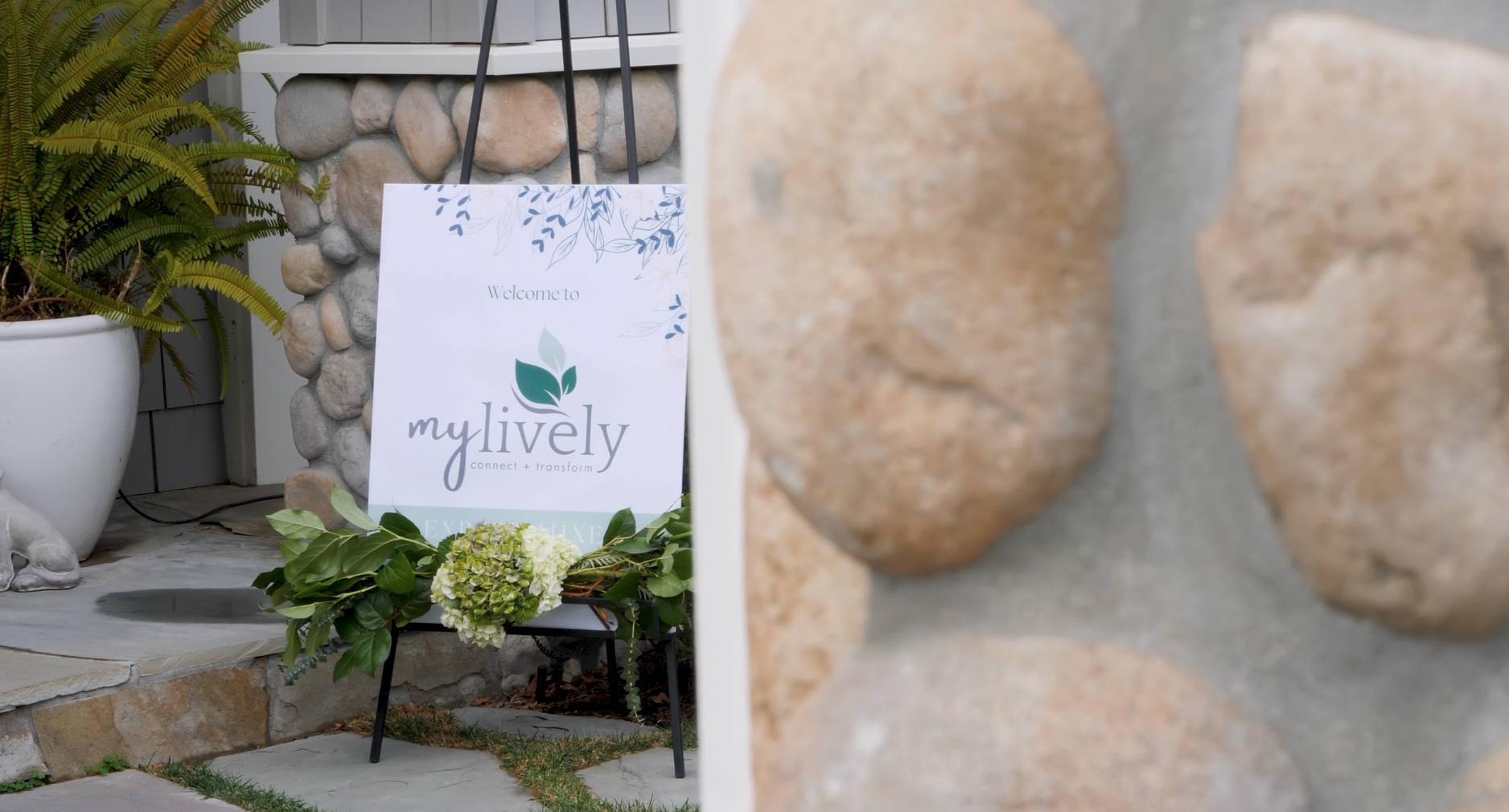 Sign reading 'Welcome to my lively, connect + transform' with greenery and stone decor.