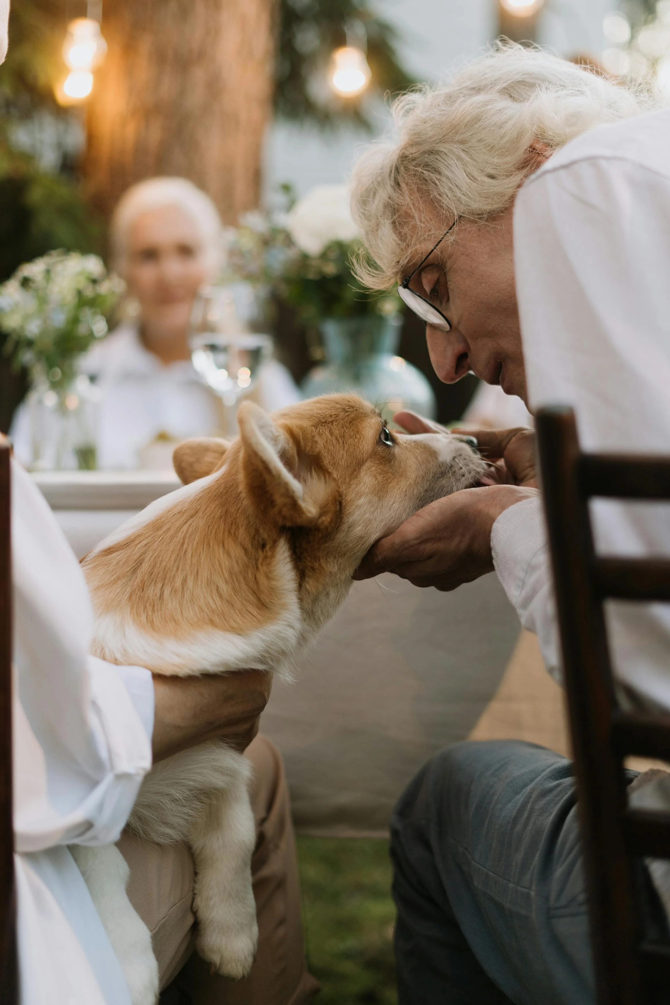 An elderly man and a young person with a puppy at a table during an outdoor gathering. The man and puppy are touching noses, with the man gently holding the puppy's face.