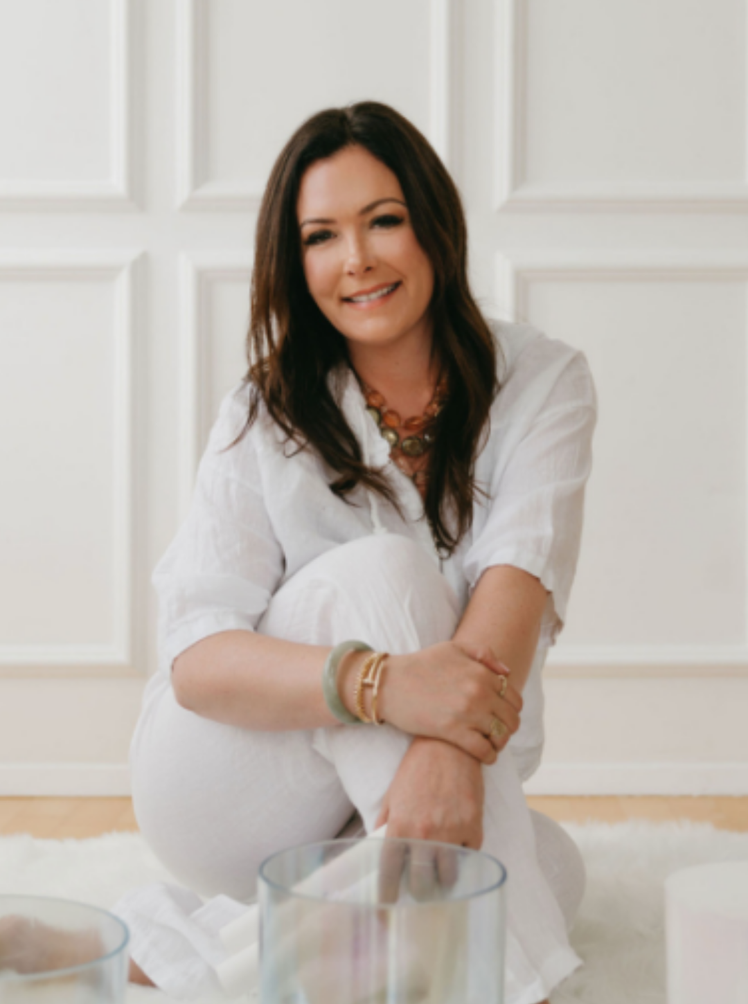 A woman with dark brown hair smiling, seated on the floor in a white outfit, surrounded by candles, in front of a white paneled wall.