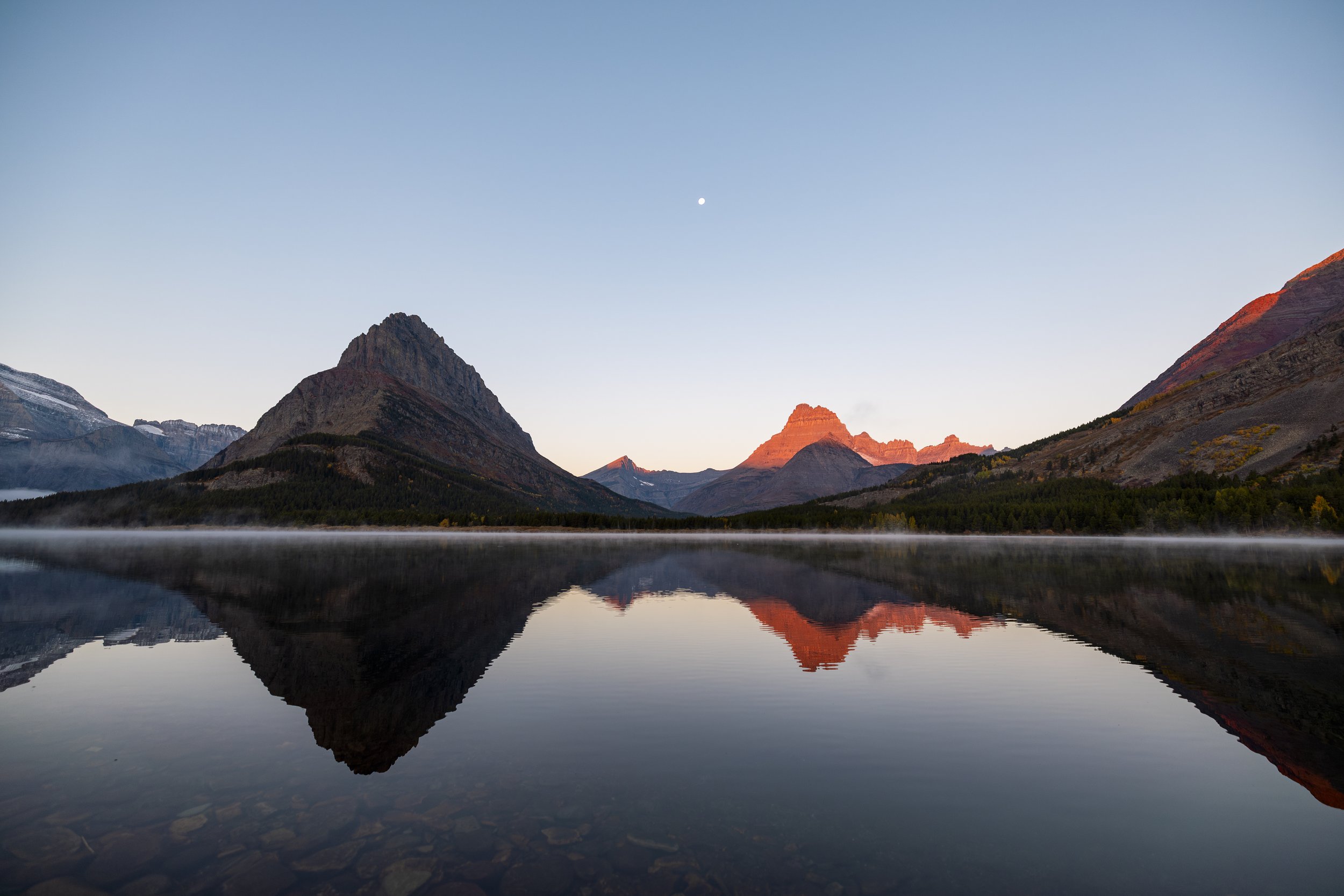 Mountain landscape with a calm lake reflecting the mountains and sky, with a visible moon in the sky.