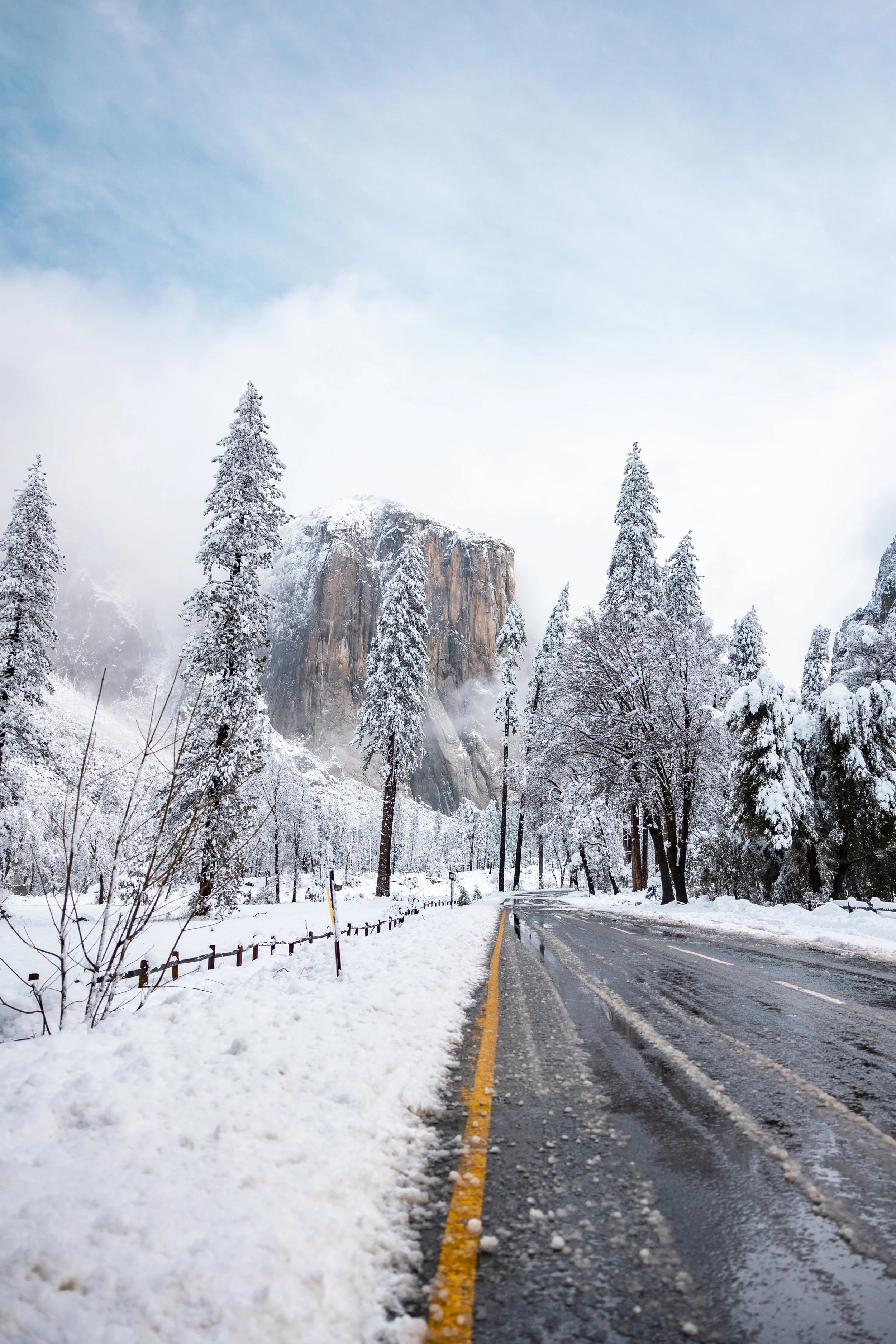 Snow-covered road winding through a forest with tall evergreen trees, leading towards a mountain with a rocky face, under a partly cloudy sky.