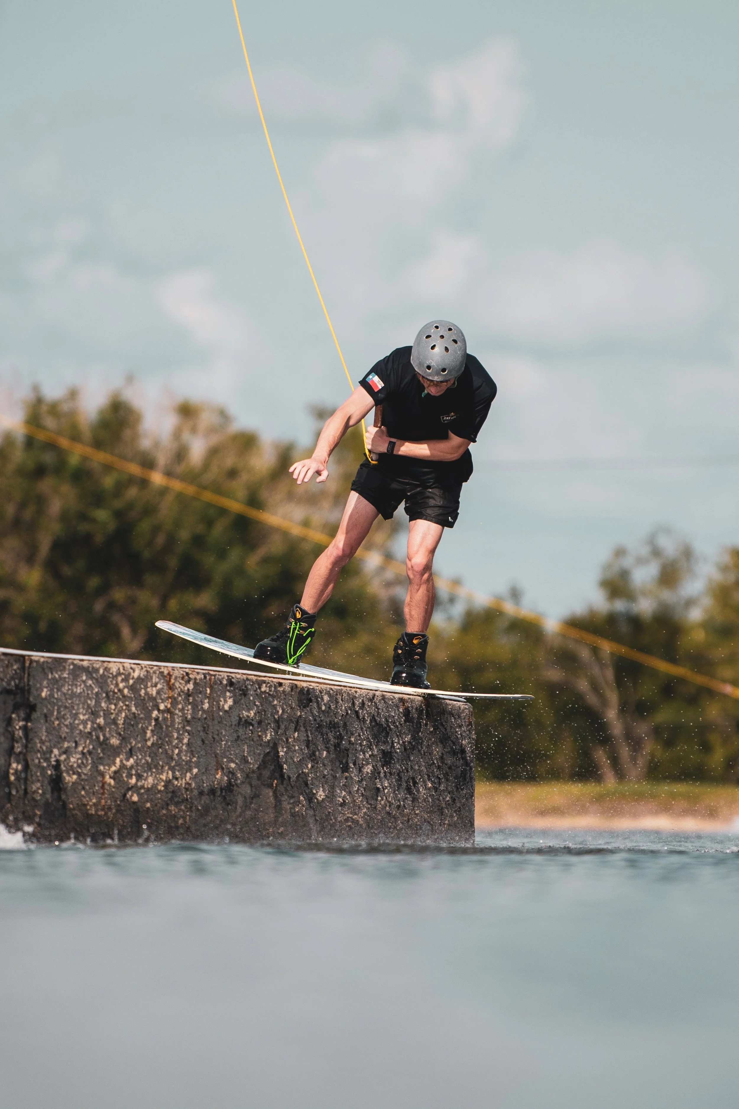 Person wearing a helmet and black clothing gliding on a skateboard on the edge of a concrete ledge over water with trees in the background.