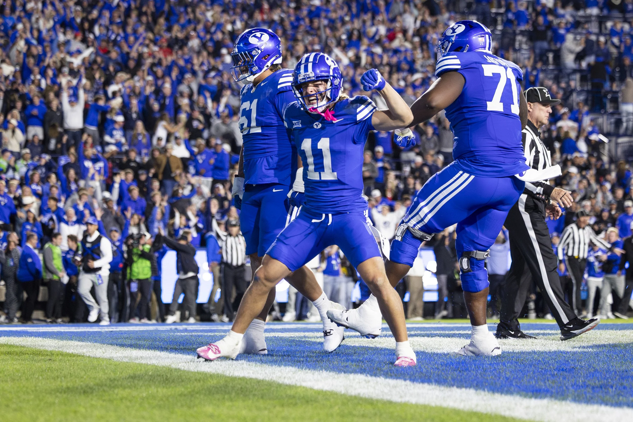 Three football players in blue jerseys celebrating on the field, with a crowd of spectators in the background.