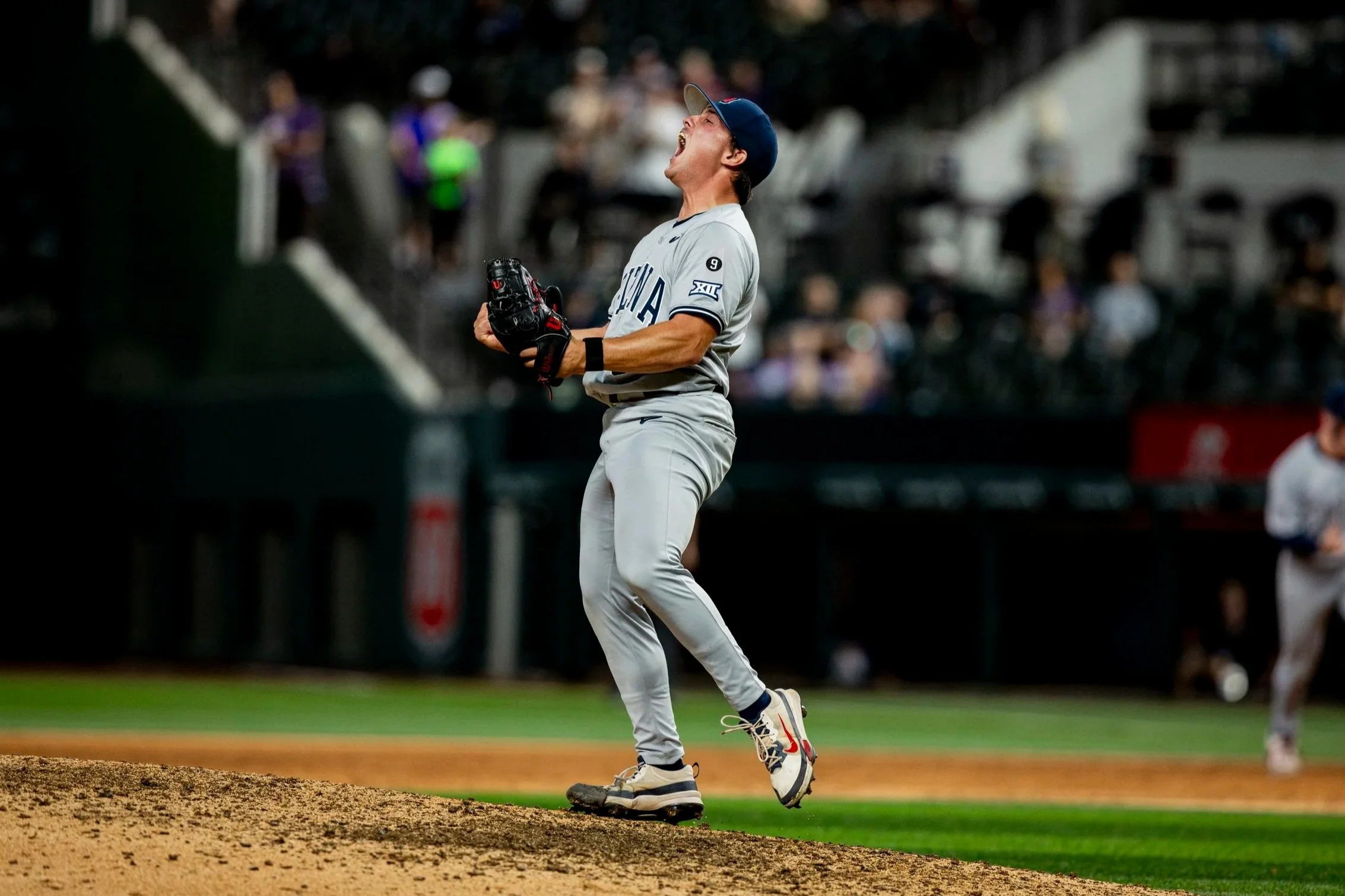 A baseball player wearing a gray uniform and a dark cap is celebrating on the pitcher's mound with his eyes closed and mouth open in excitement, holding a glove in his left hand. Other players and spectators are visible in the background.