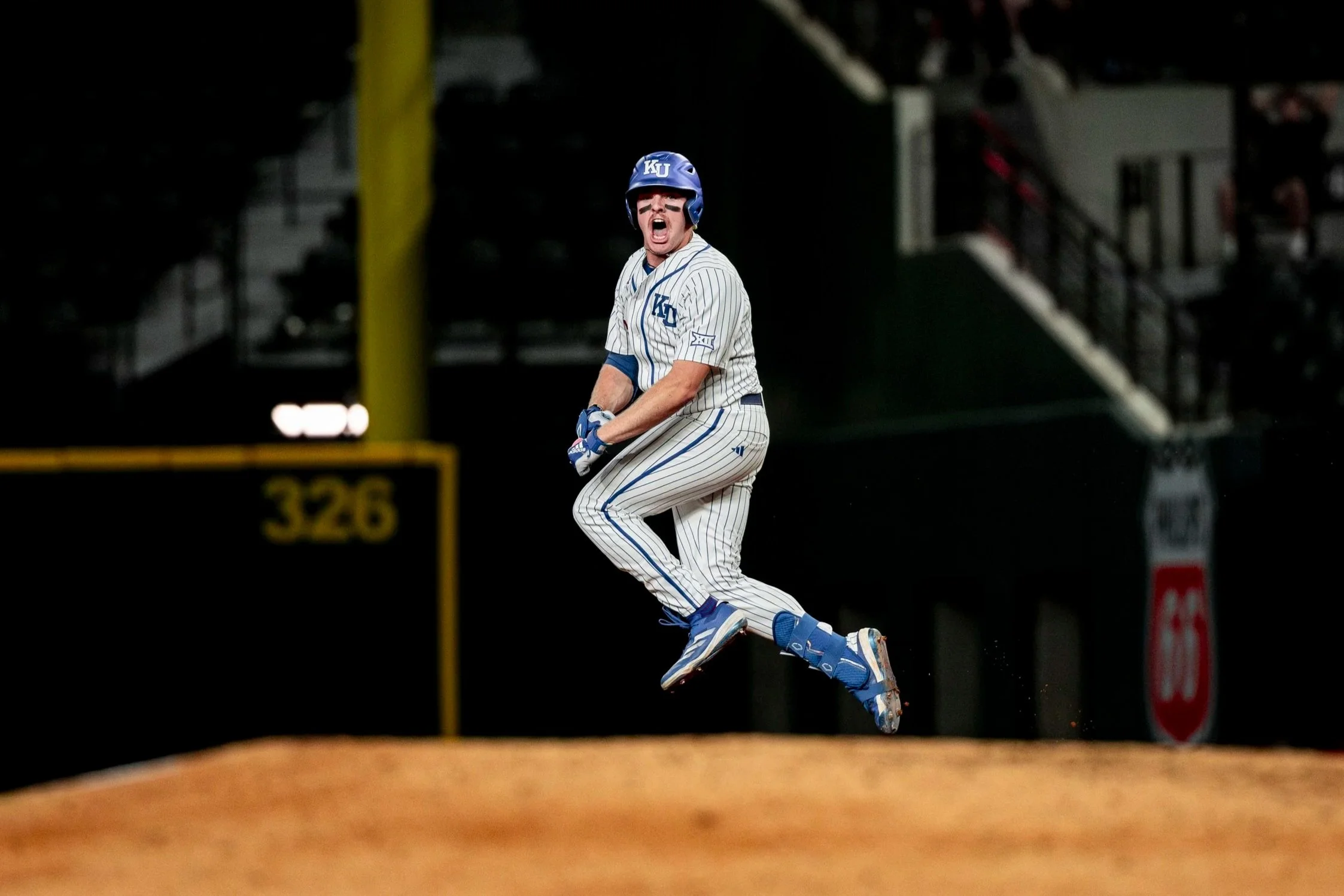 A baseball player in a white pinstripe uniform jumping in the air with an excited expression, wearing a blue helmet and gear, on a baseball field at night.