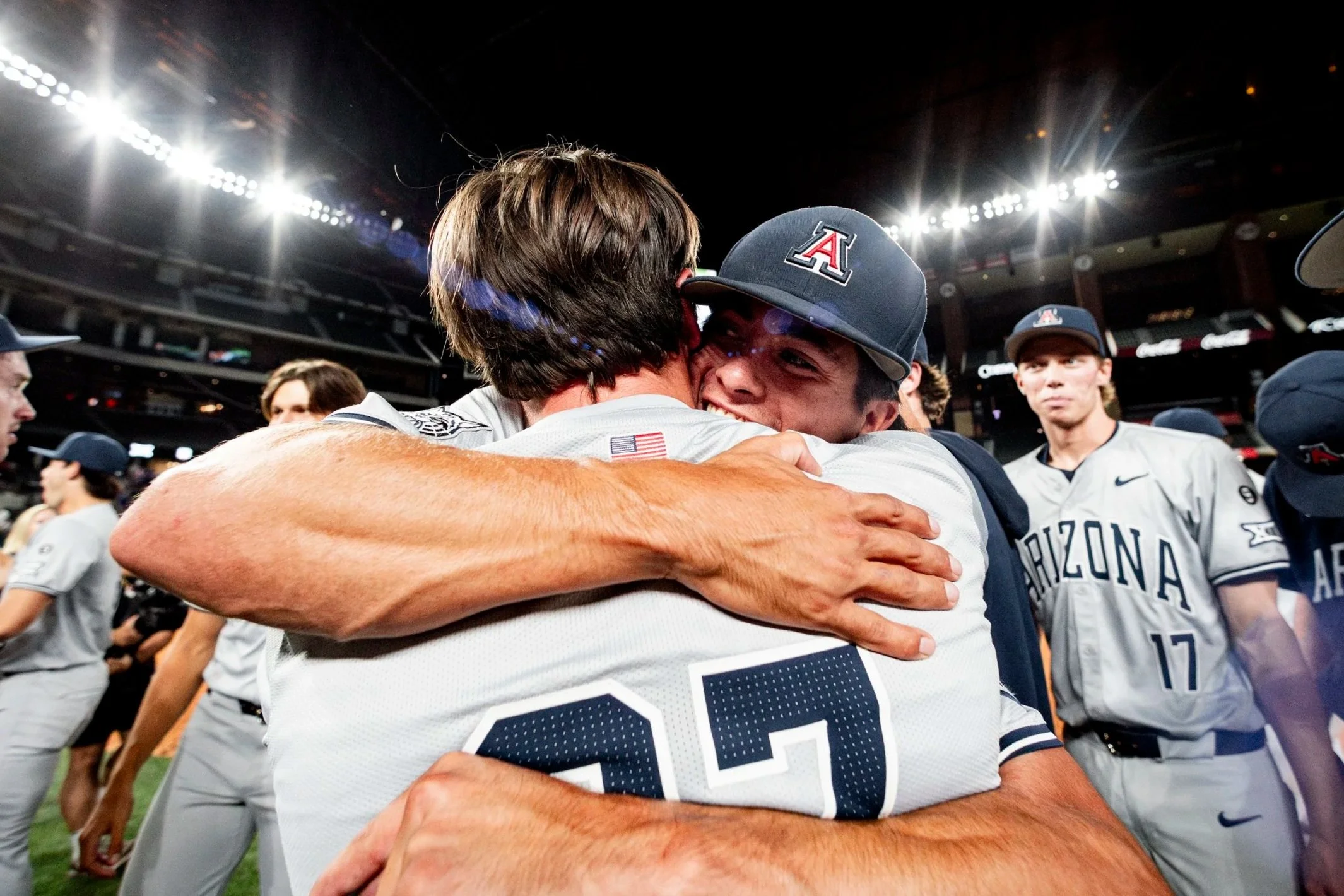 Two baseball players from Arizona Wildcats team hugging on the field after a game, surrounded by teammates under stadium lights.