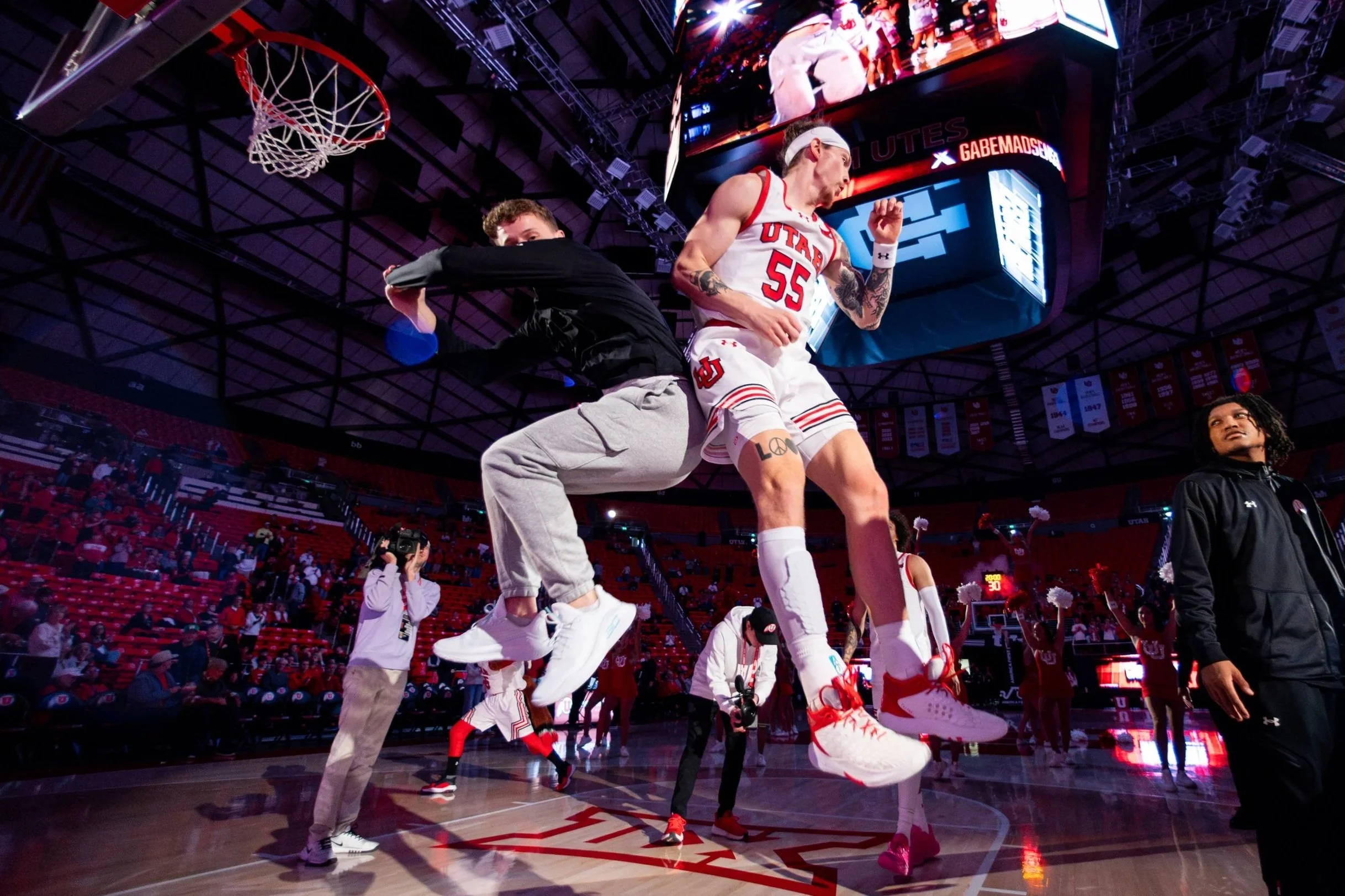 Two basketball players mid-air during pregame warm-up, with a basketball hoop overhead and cheerleaders in the background on the court.
