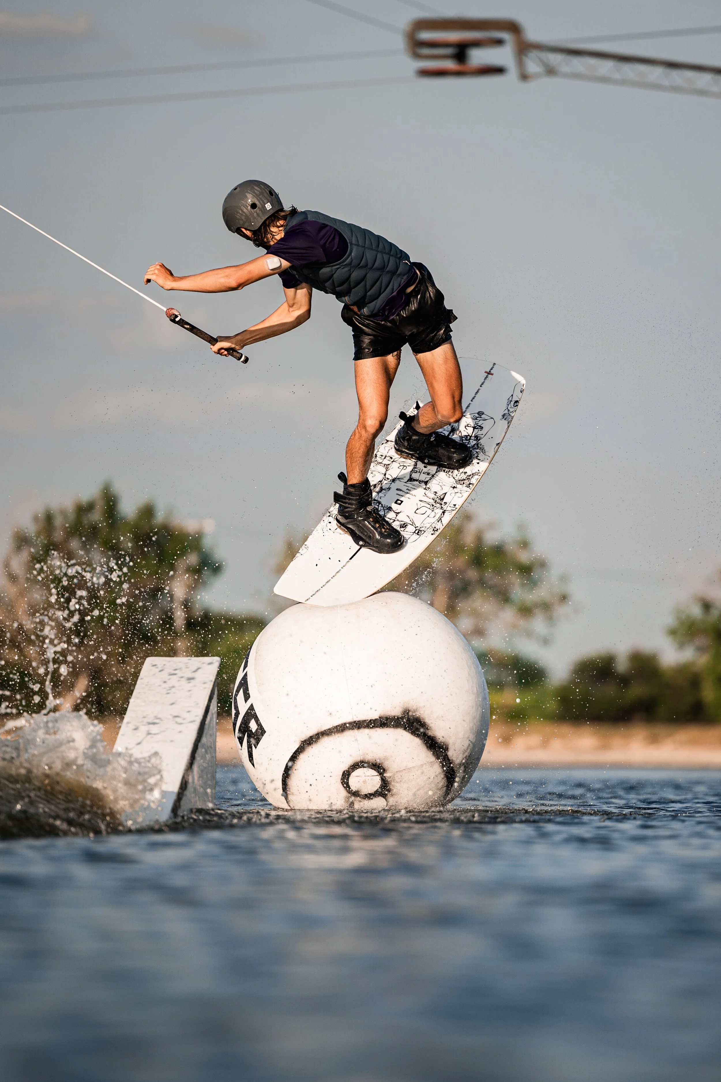 A person wakeboarding on water, performing a jump over a large buoy with a spherical shape, wearing a helmet, life vest, and shorts.