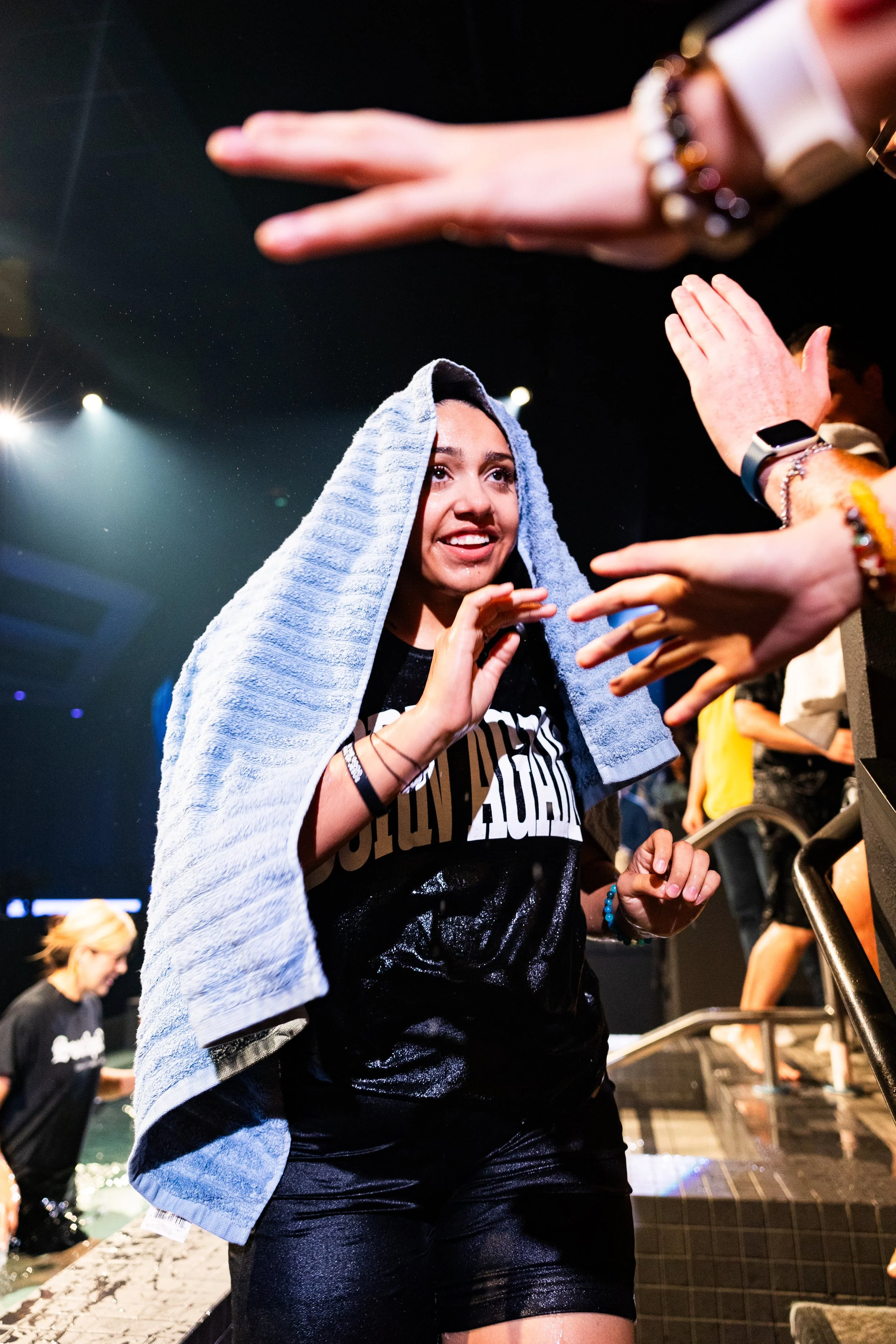 Young woman with a towel on her head smiling as she receives a blessing or prayer from people with outstretched hands in a dark indoor setting.