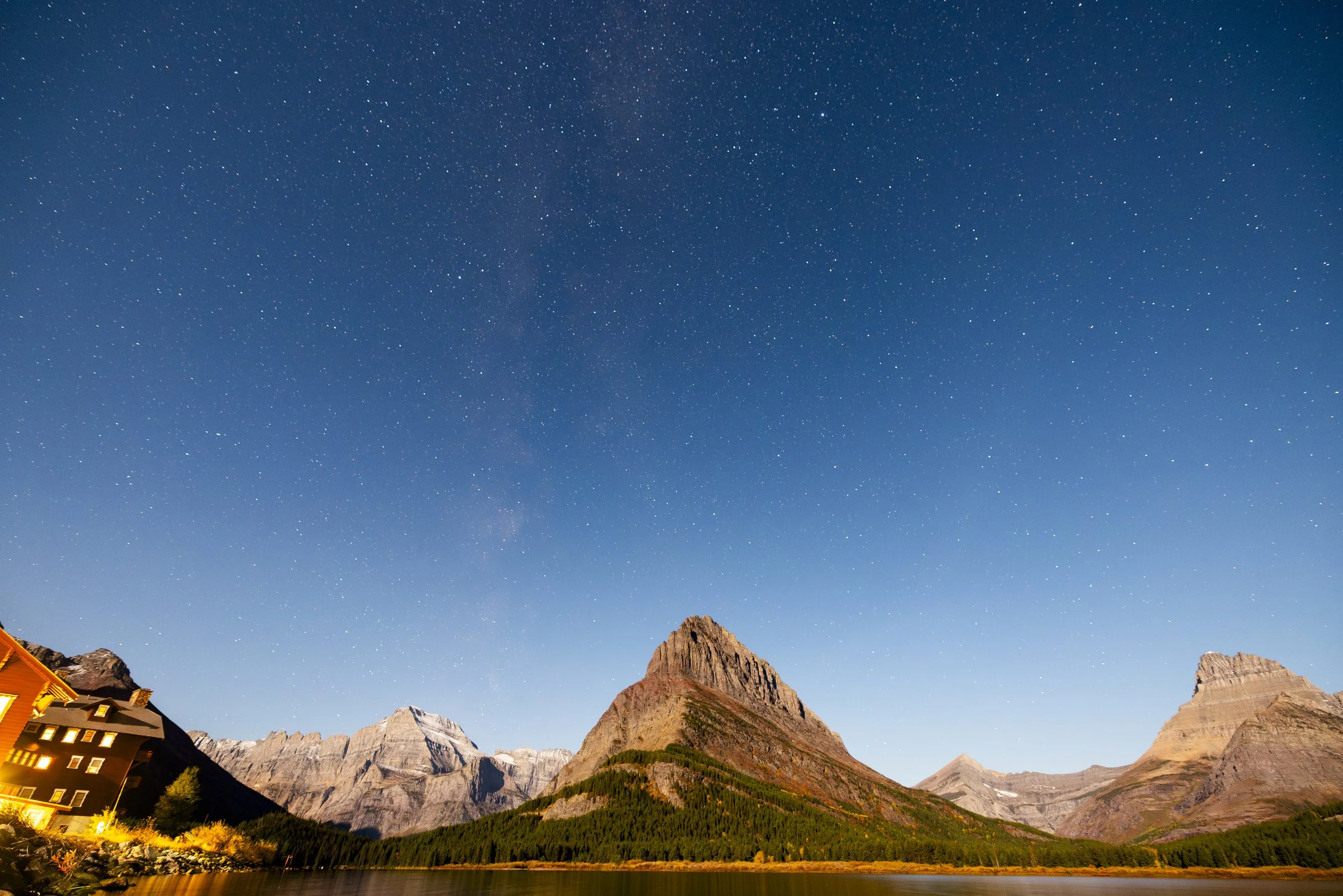 Nighttime view of a mountain landscape with a clear starry sky and a small lit building on the left.