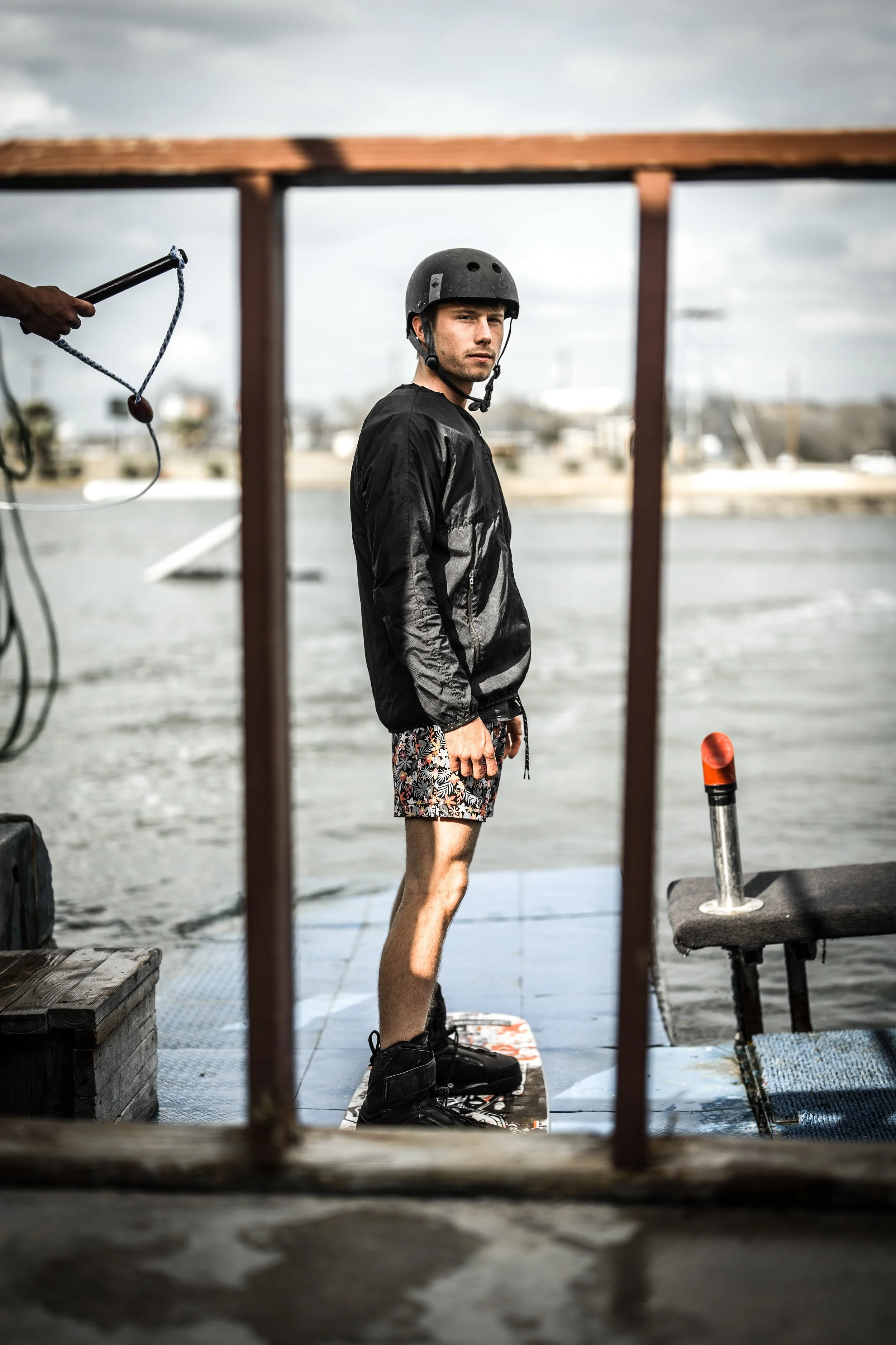A young man wearing a black helmet, black jacket, patterned shorts, and black water shoes standing on a wakeboarding boat deck, looking into the distance as a person holds a rope nearby, with water and cloudy sky in the background.