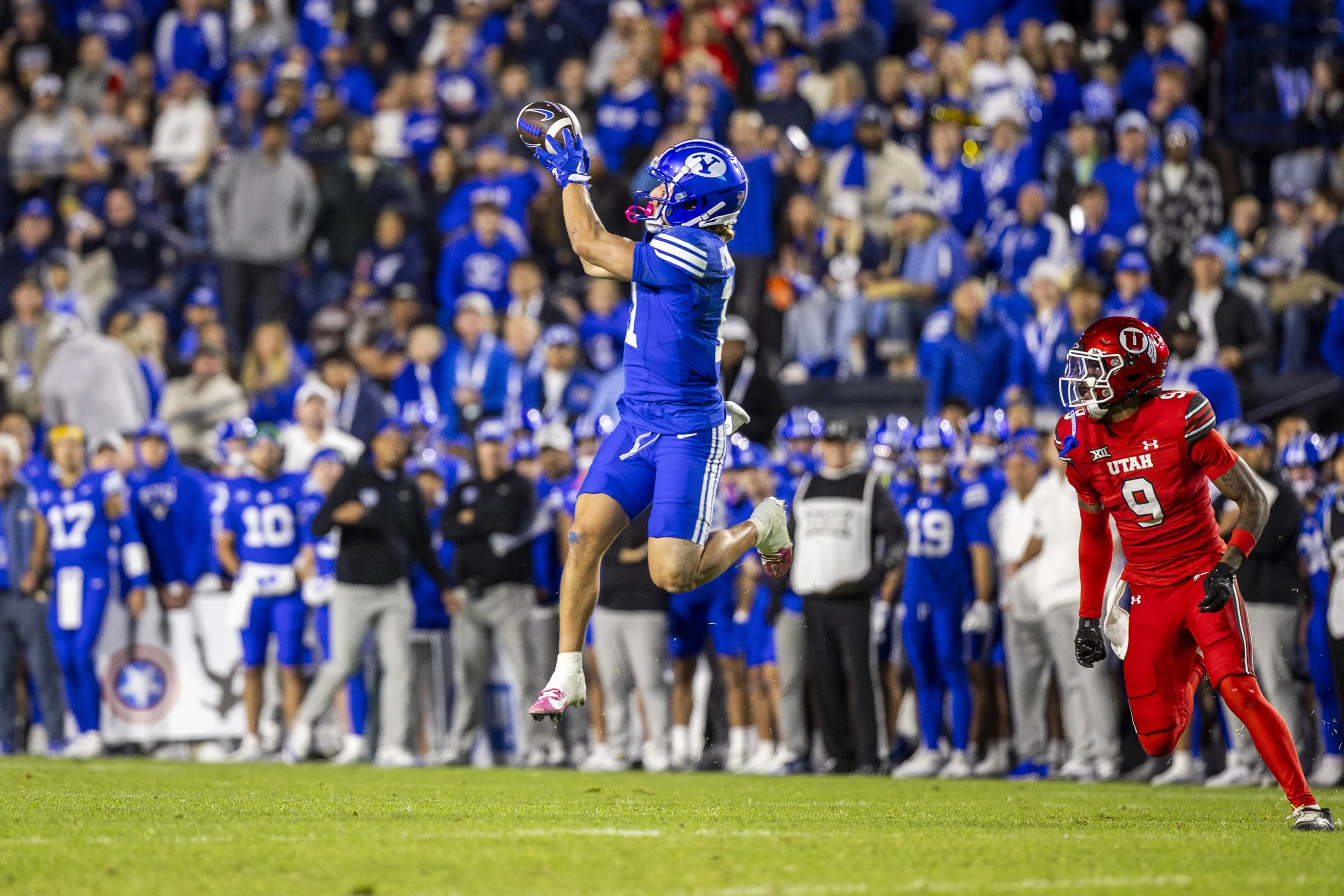 A football player in a blue uniform jumps to catch a football during a game. An opposing player in a red uniform stands nearby. The crowd in the background is dressed in blue and watching the game.
