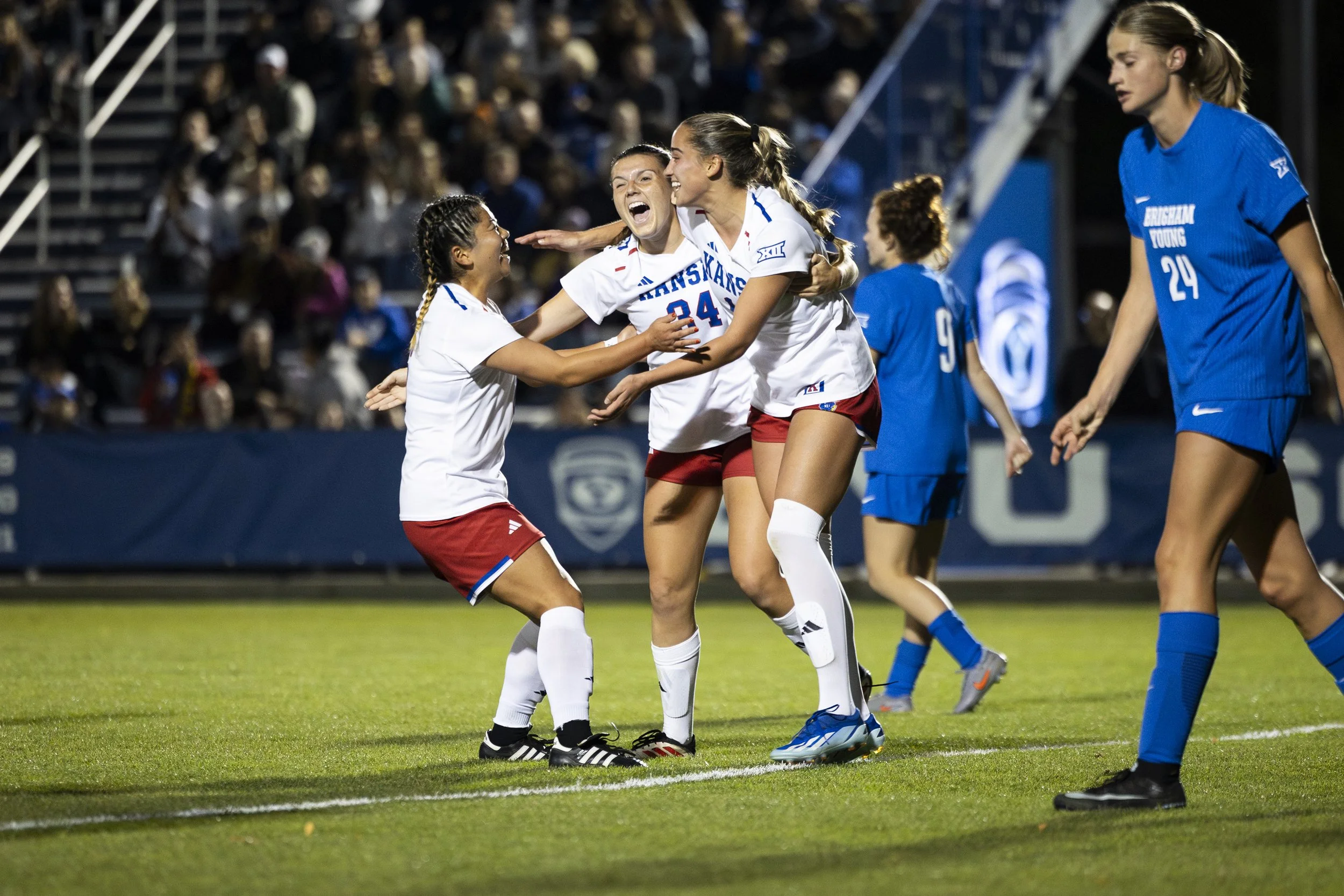 Soccer players celebrating on the field during a match, with one player in red shorts and white jersey hugging a teammate, and others in the background under stadium lights.