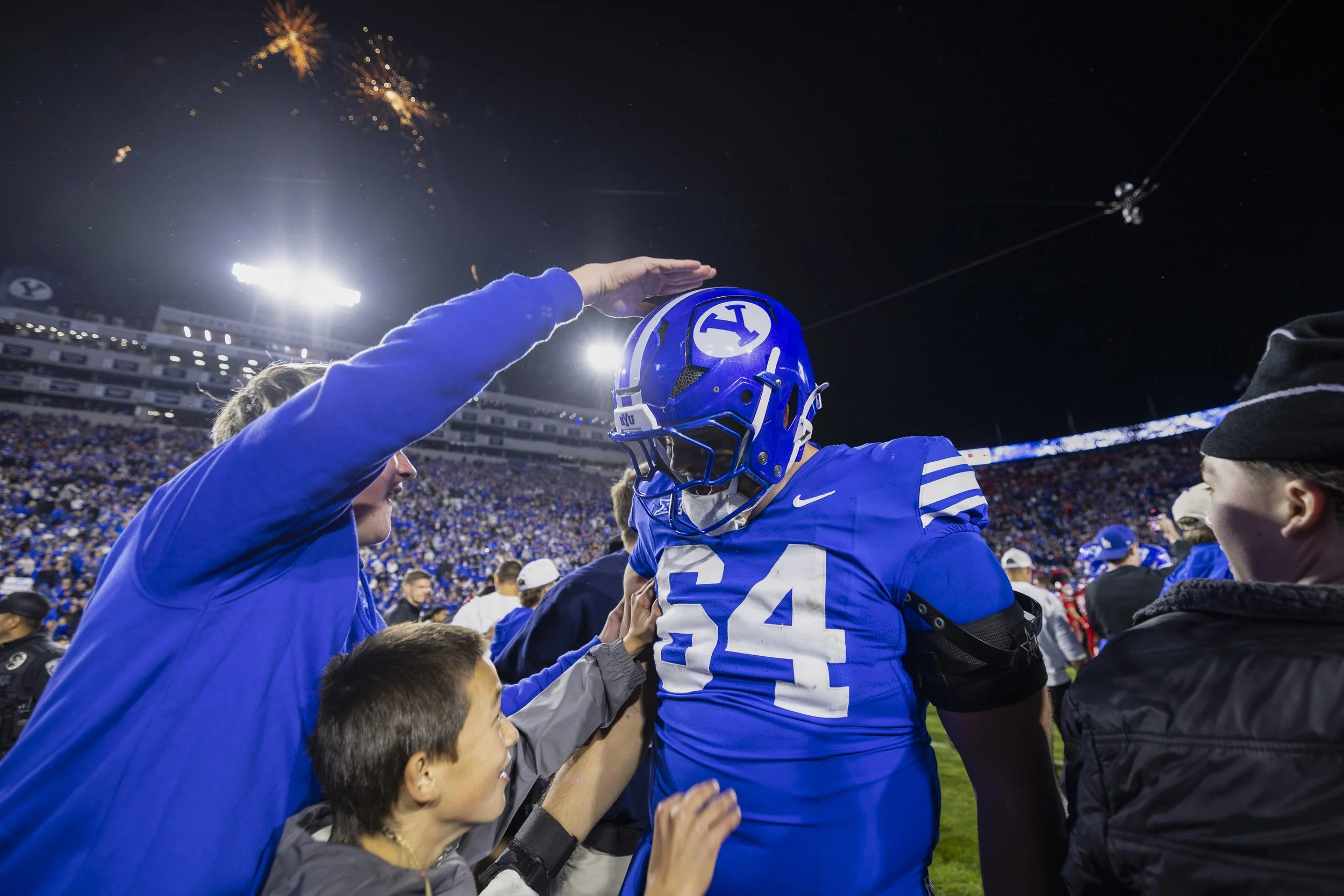 A football player wearing a blue uniform with the number 54 is surrounded by fans at a stadium. One fan is touching his helmet, and a boy is reaching out to him. The stadium is filled with spectators, and fireworks are visible in the night sky.