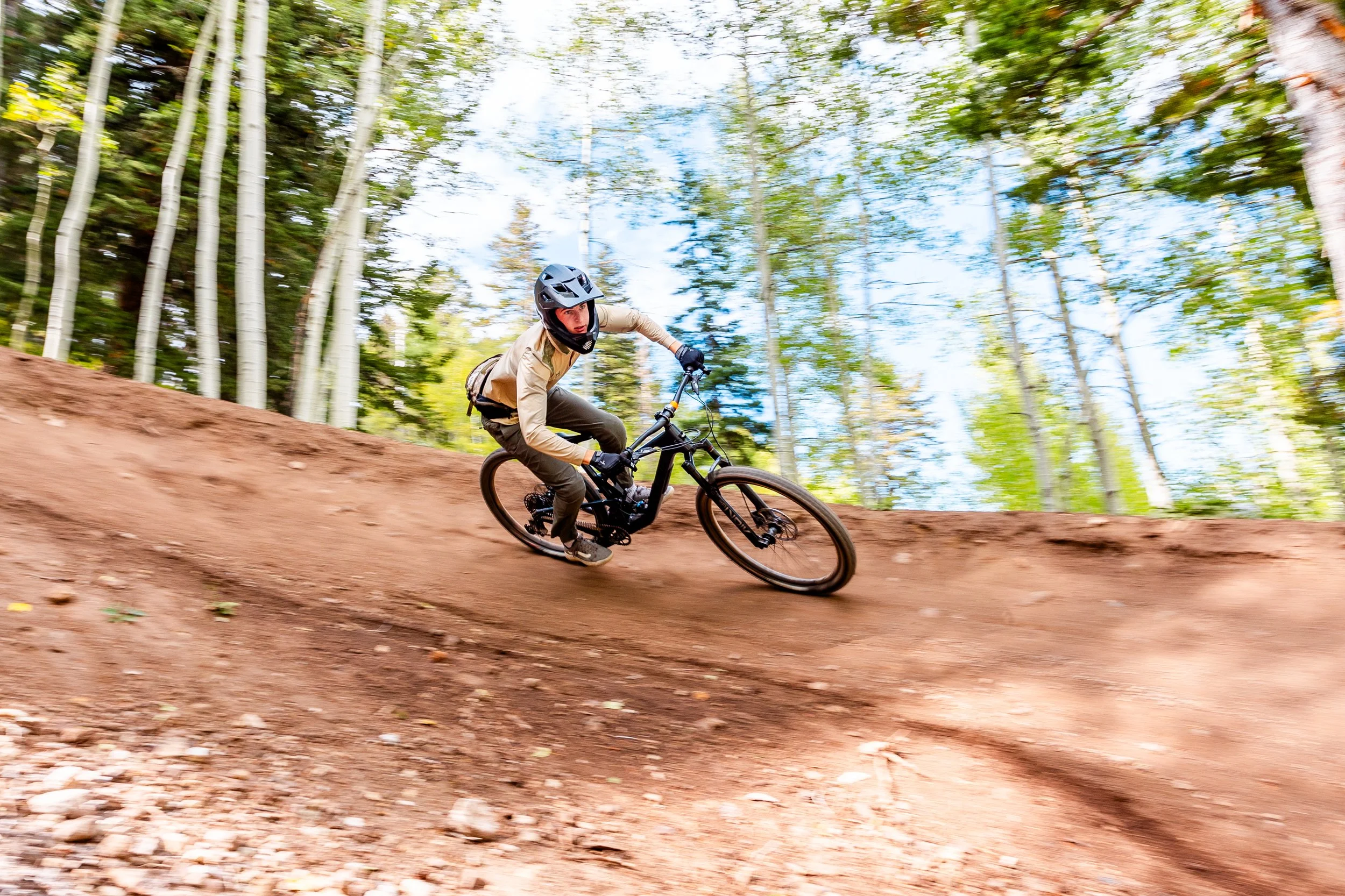 A person mountain biking on a dirt trail in a forest with trees and blue sky in the background.