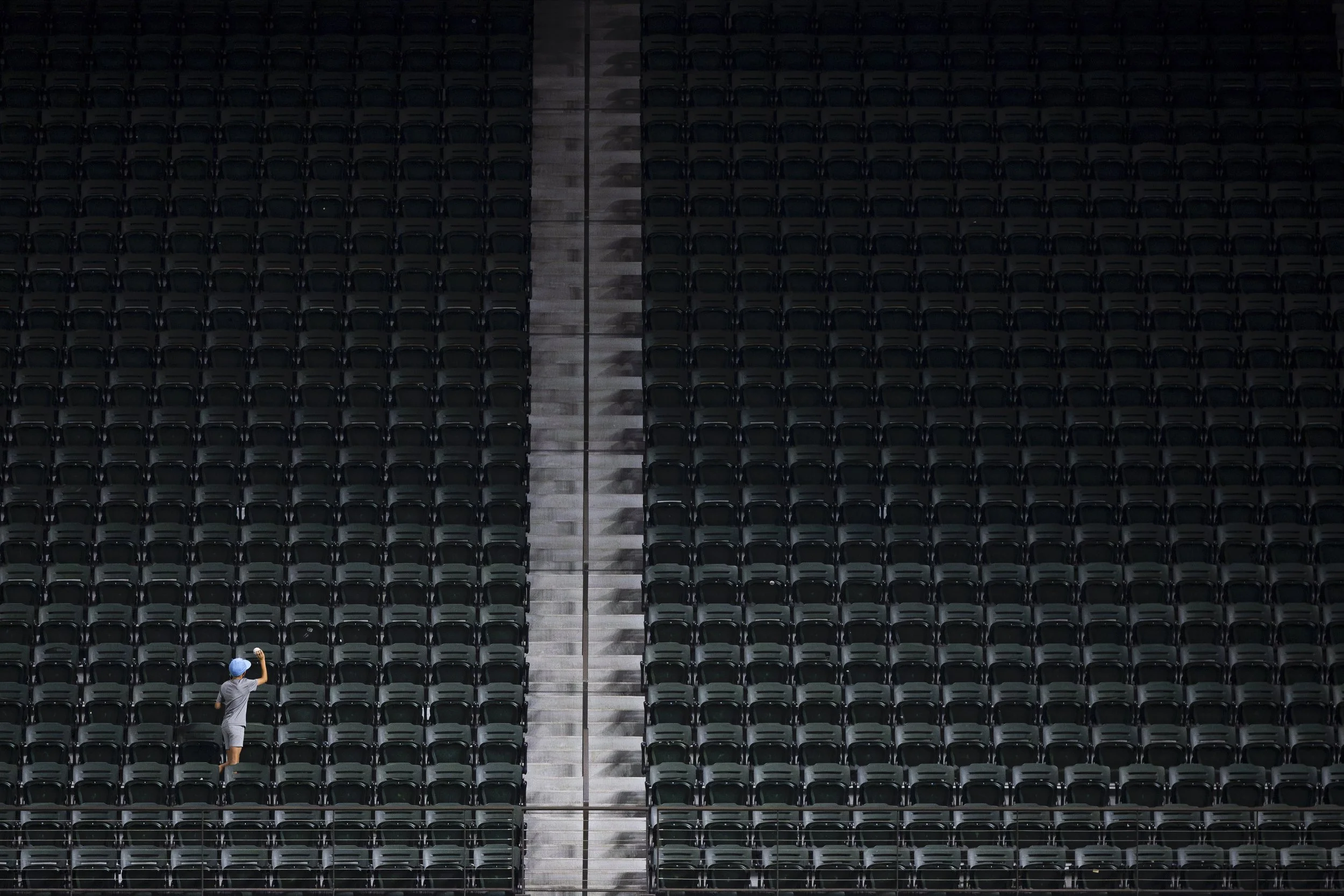 A person wearing a gray shirt, shorts, and a blue cap standing among empty black stadium seats with a walkable aisle in between.