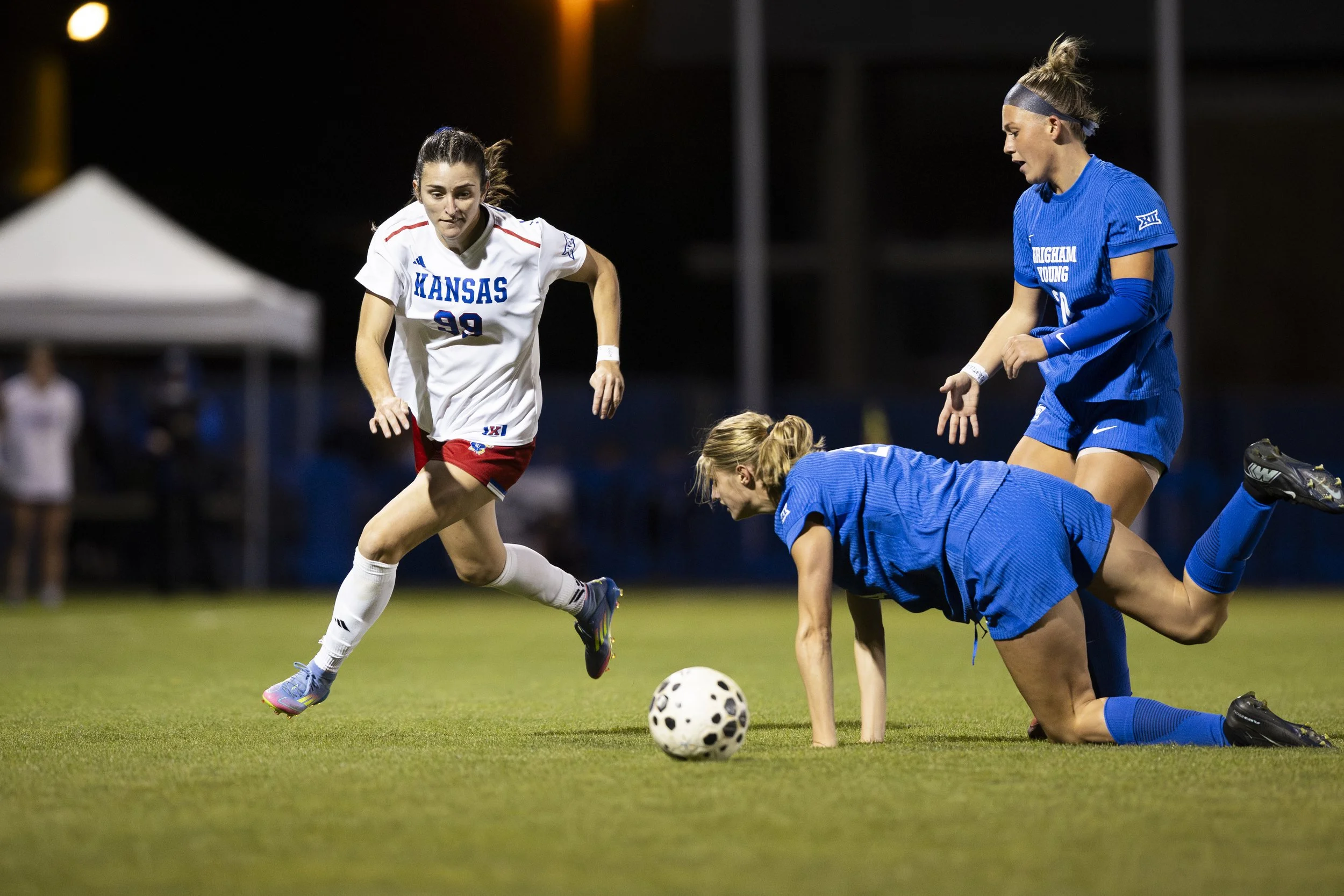 Women's soccer match with four players, two in blue uniforms and two in white and red uniforms, on a grassy field at night.