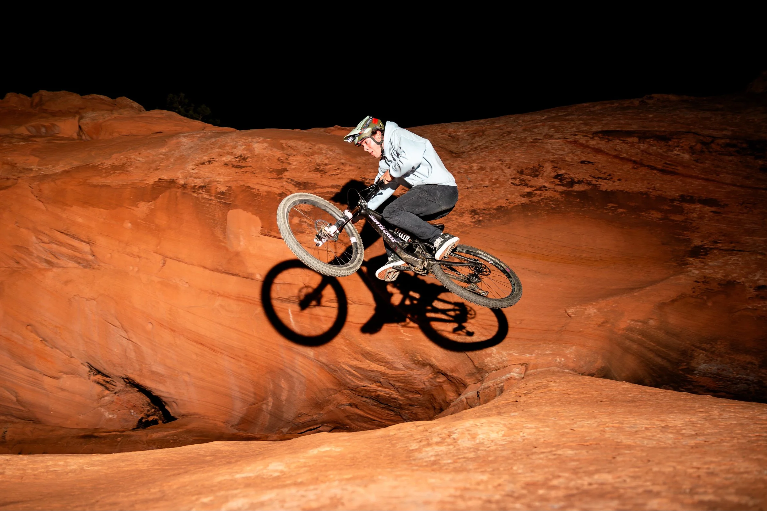 Person wearing helmet and light jacket riding mountain bike on red rock terrain at night, illuminated with bright light, creating a distinct shadow.