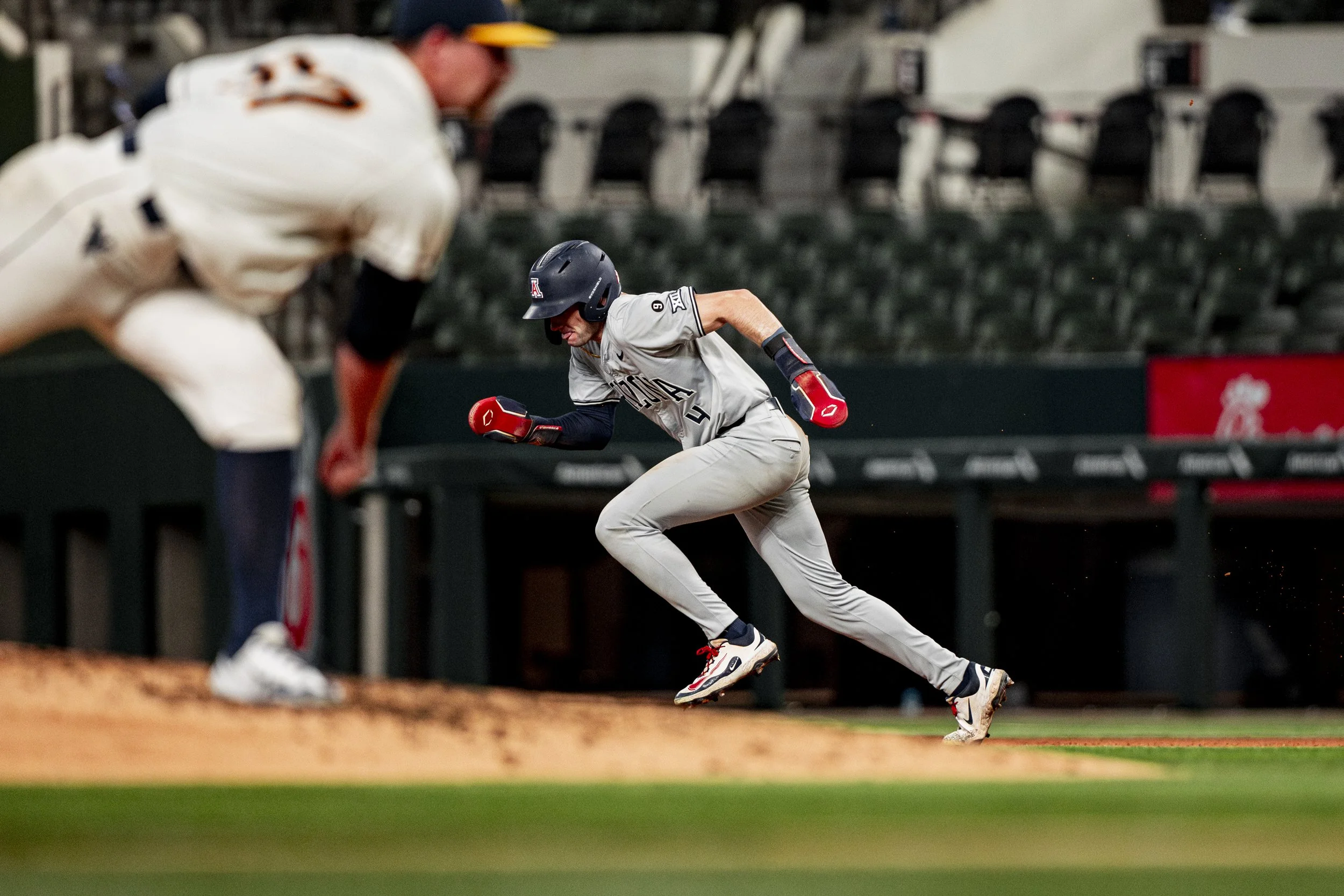 A baseball player from Arizona is sprinting towards a base during a game, wearing a helmet, gray uniform, and red gloves, with the catcher's blurred figure in the foreground.