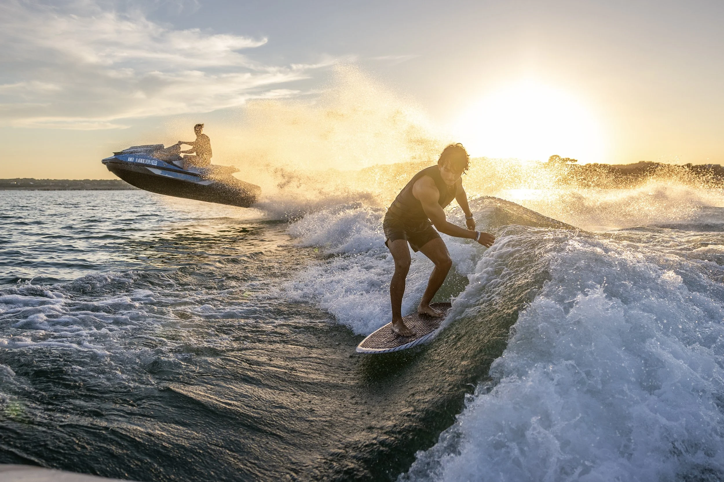 Two people riding jet skis on the water during sunset, with one person surfing on a wave in the foreground.