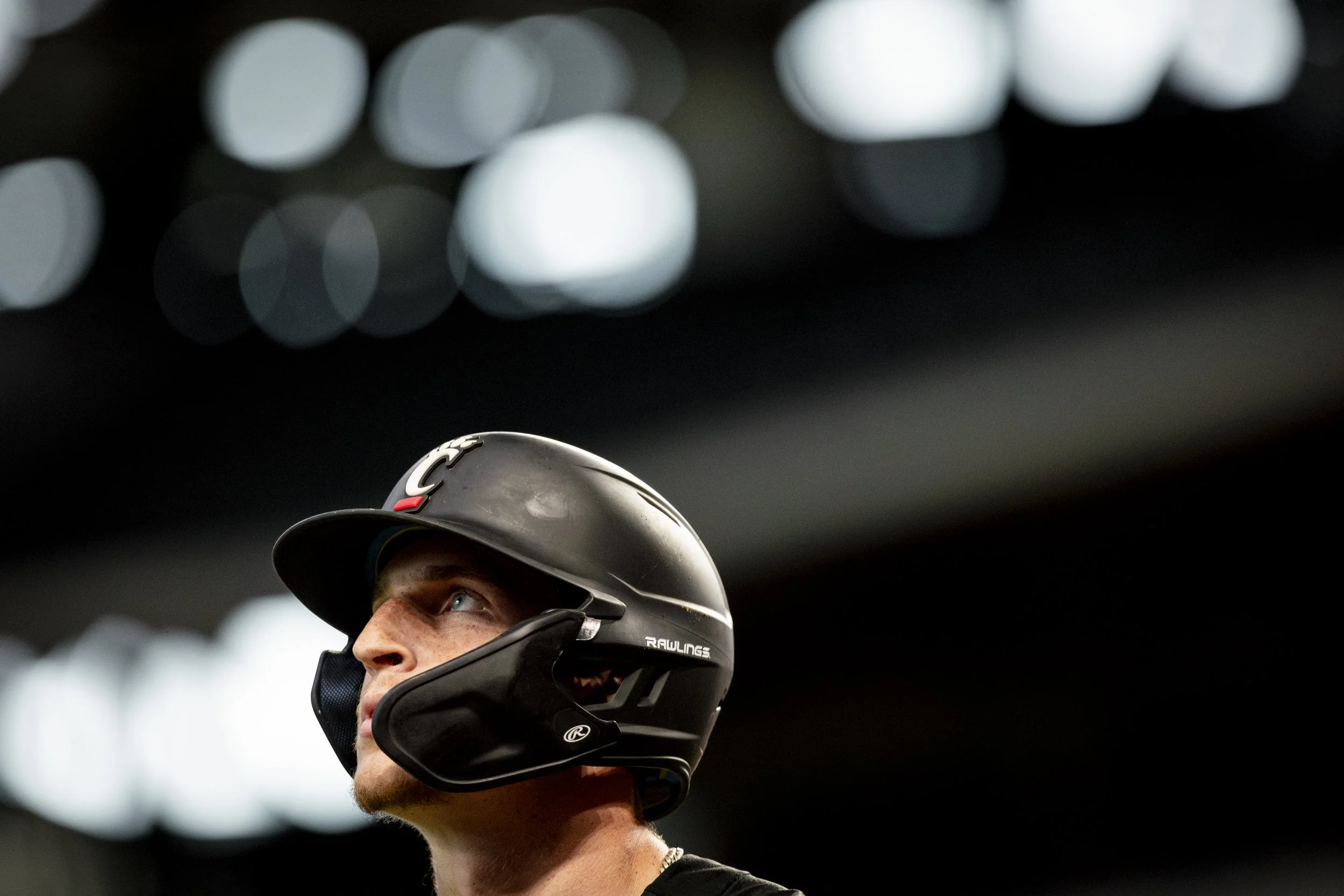A man wearing a black baseball helmet with a face guard, looking upwards inside a stadium with bright lights in the background.