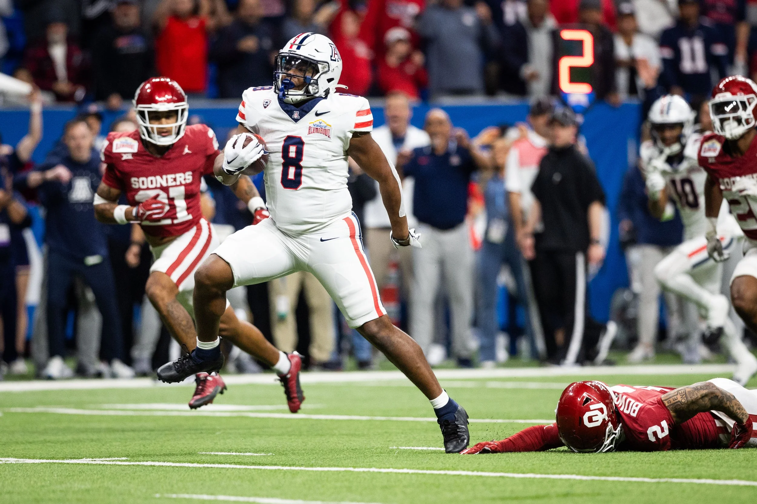 American football game with player in white uniform running with the ball, players in red and white uniforms on the field, and a crowd in the background.