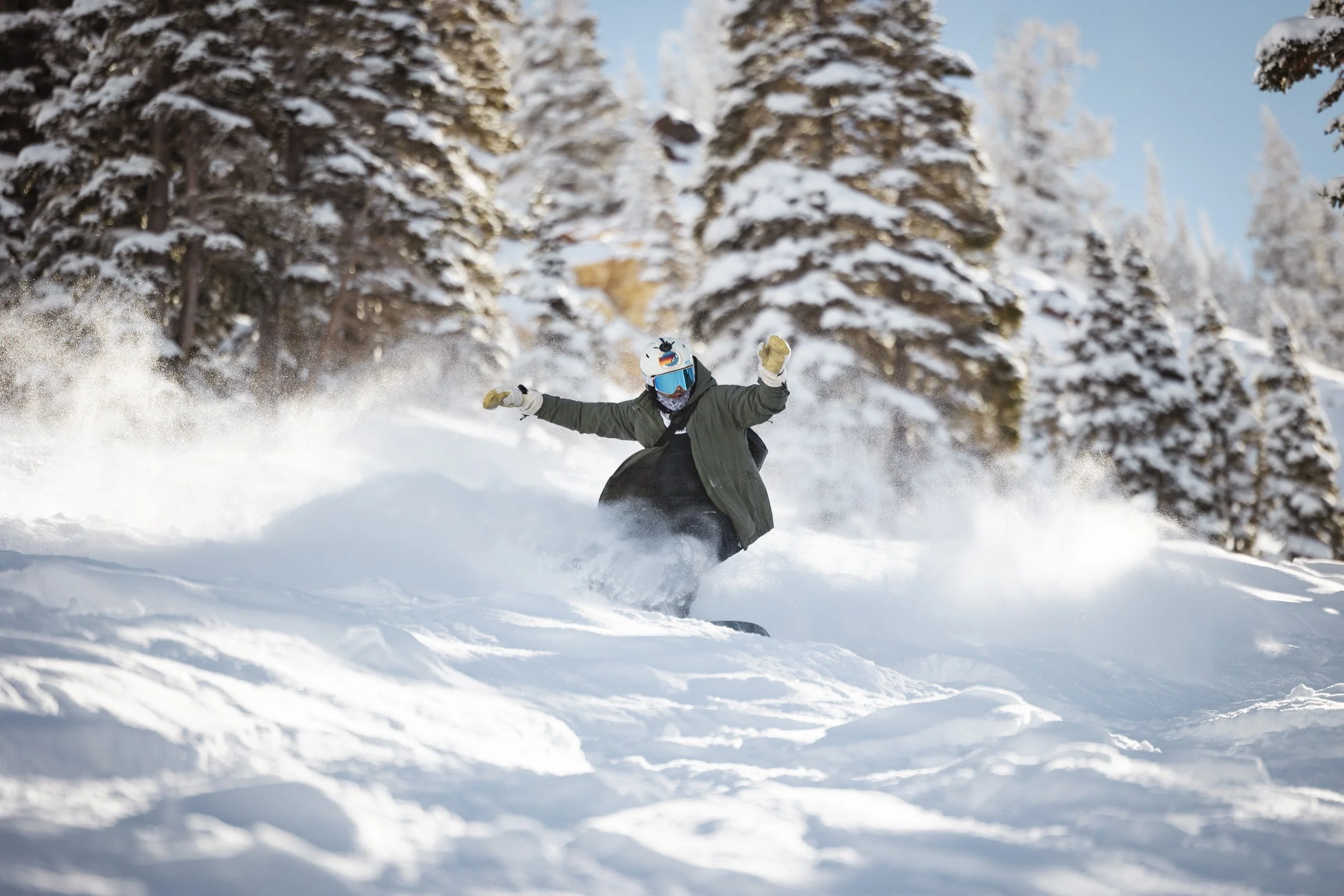 Person snowboarding down a snowy slope surrounded by snow-covered trees, wearing a helmet, goggles, and winter gear.