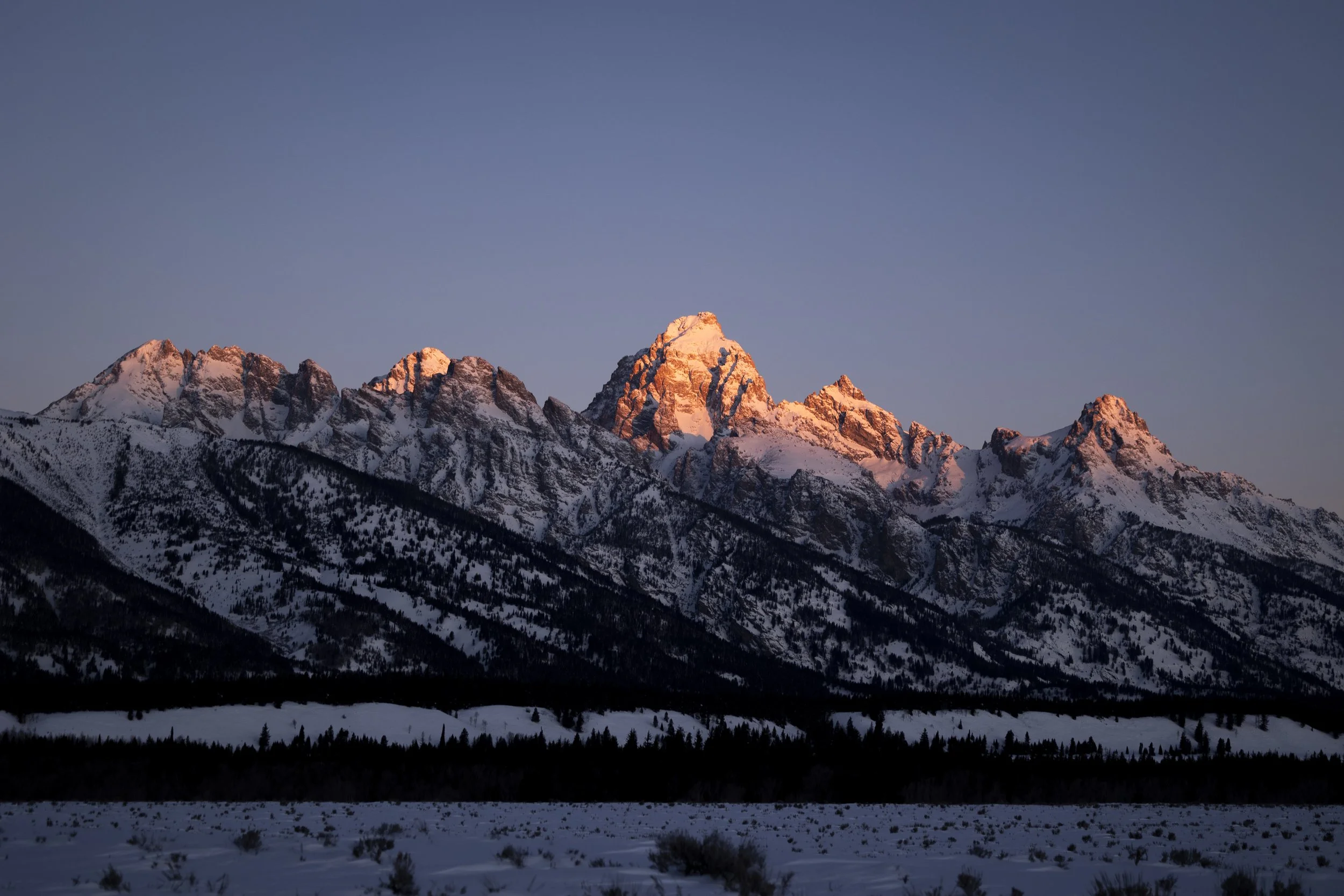 Snow-covered mountain range at sunset, with peaks illuminated by pinkish light, under a clear blue sky.